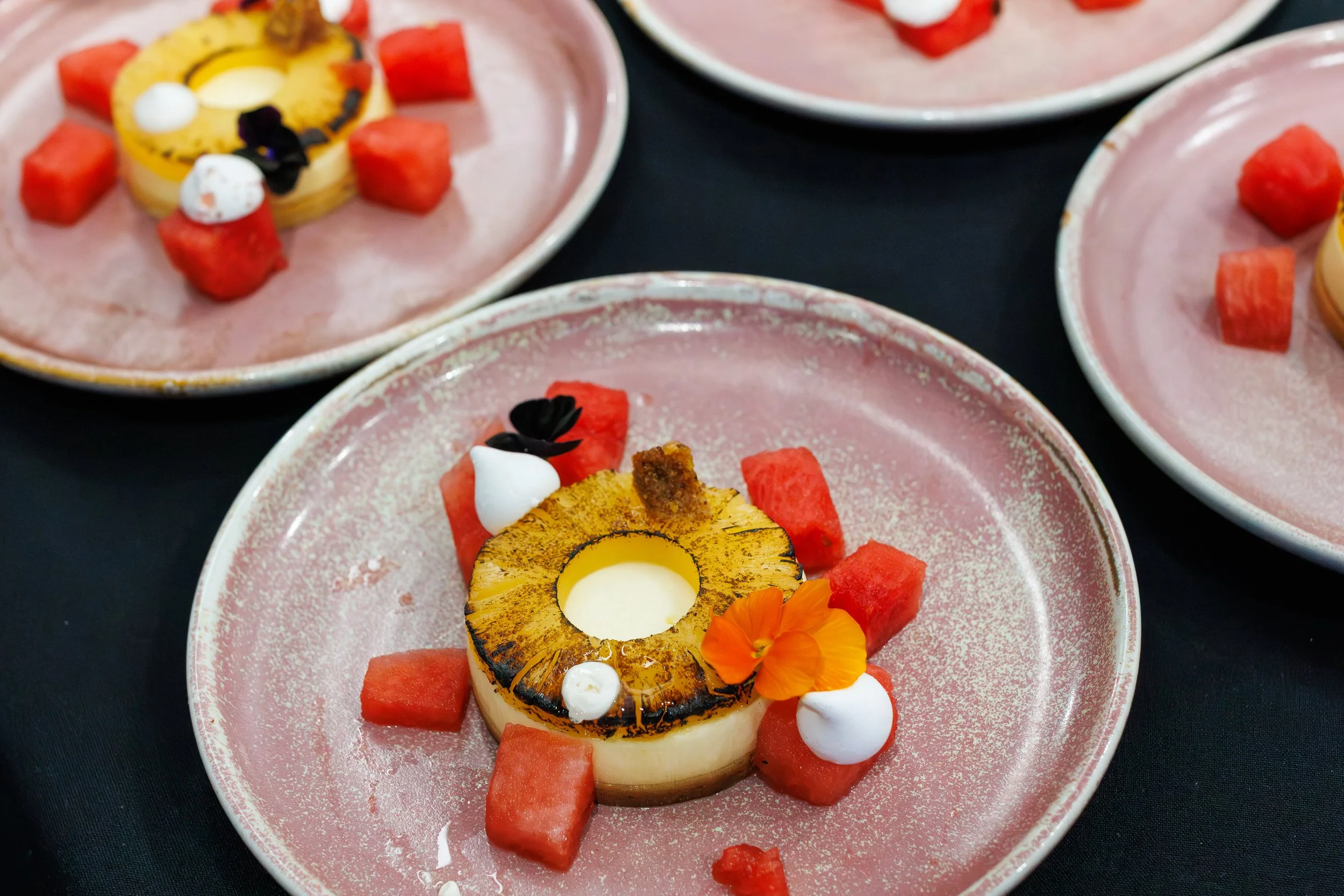 A dessert with a slice of roasted pineapple topped with white cream, surrounded by watermelon cubes, small white dollops, and orange edible flowers on a pink plate.