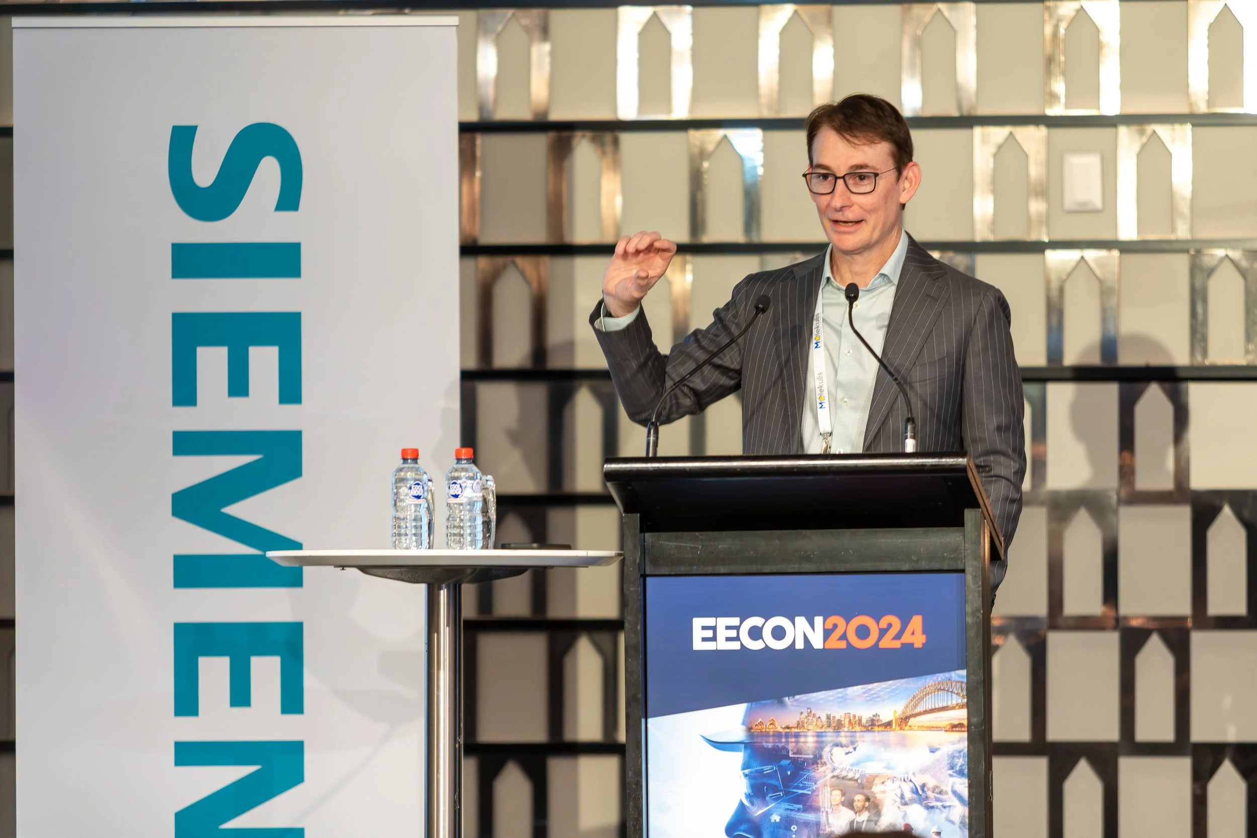 A man wearing glasses and a suit delivers a speech at a conference, with a large banner displaying the event name 'EECON 2024' and a cityscape image in the background. There are four bottles of water on a small round table beside him.