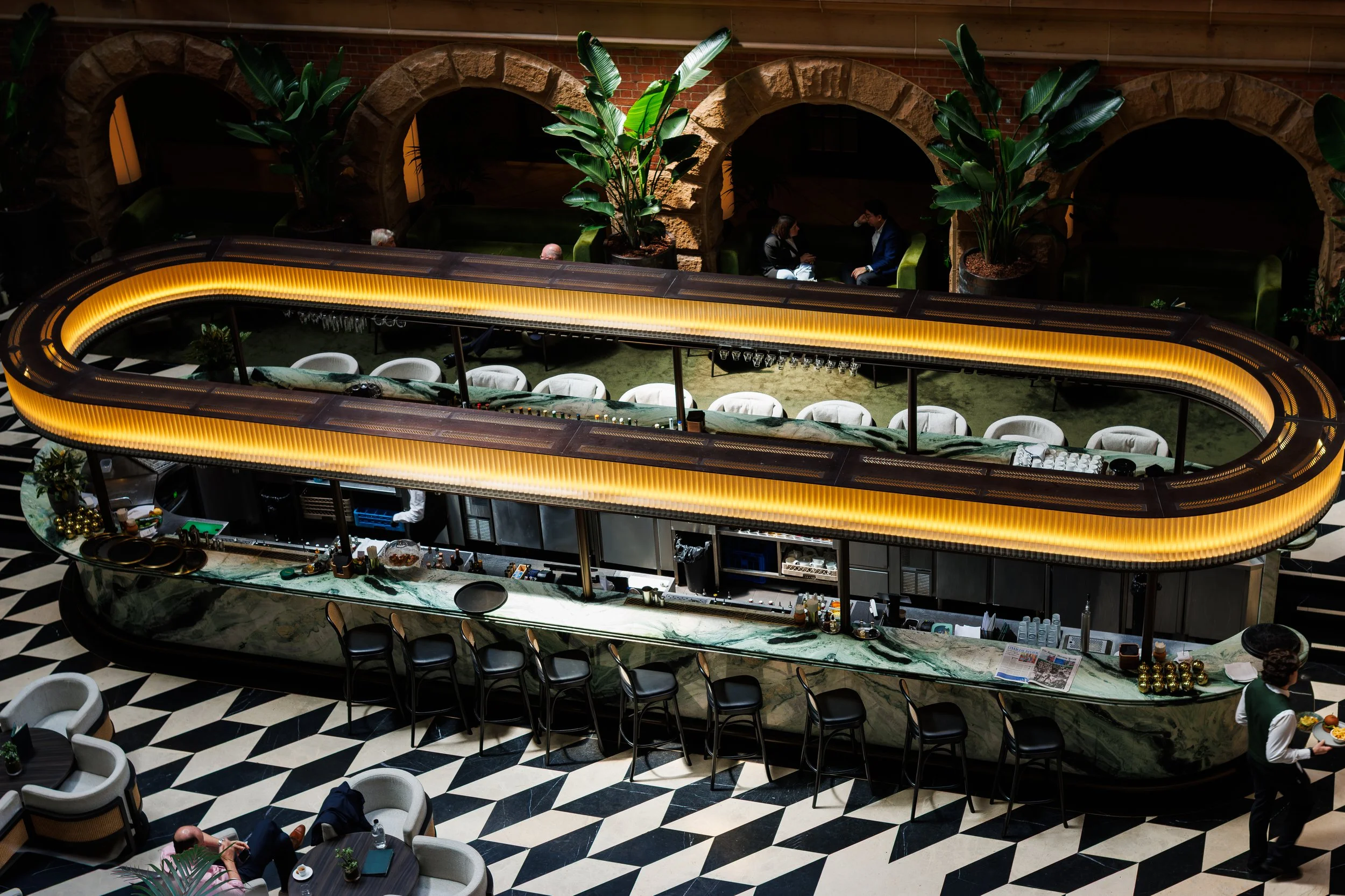 A bar area with a curved illuminated counter and white chairs, surrounded by large green plants and seating booths in the background, with people dining and chatting.
