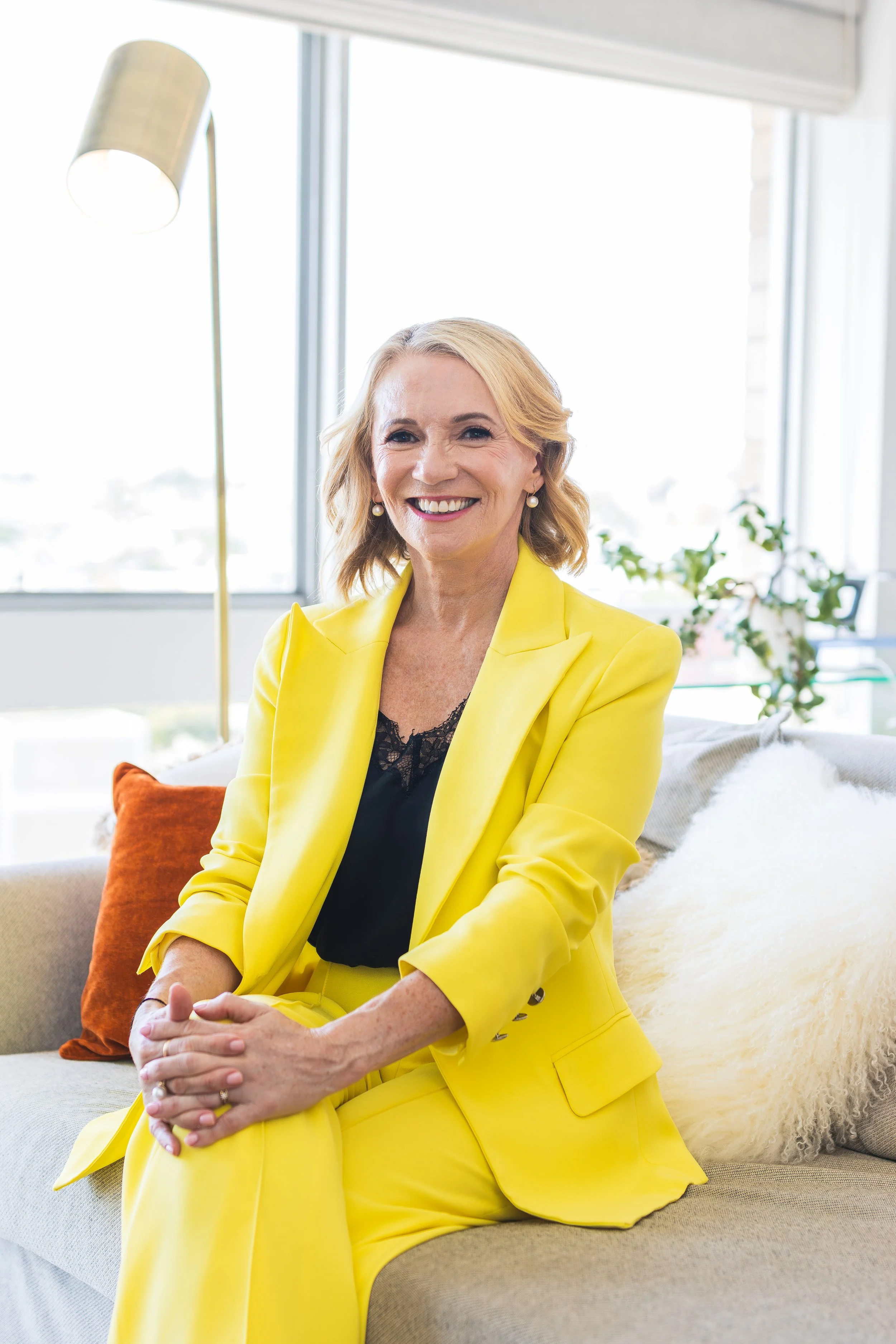 A smiling woman with blonde hair wearing a bright yellow blazer and matching pants sitting on a beige sofa in a well-lit room with large windows, a green plant, and colorful pillows.