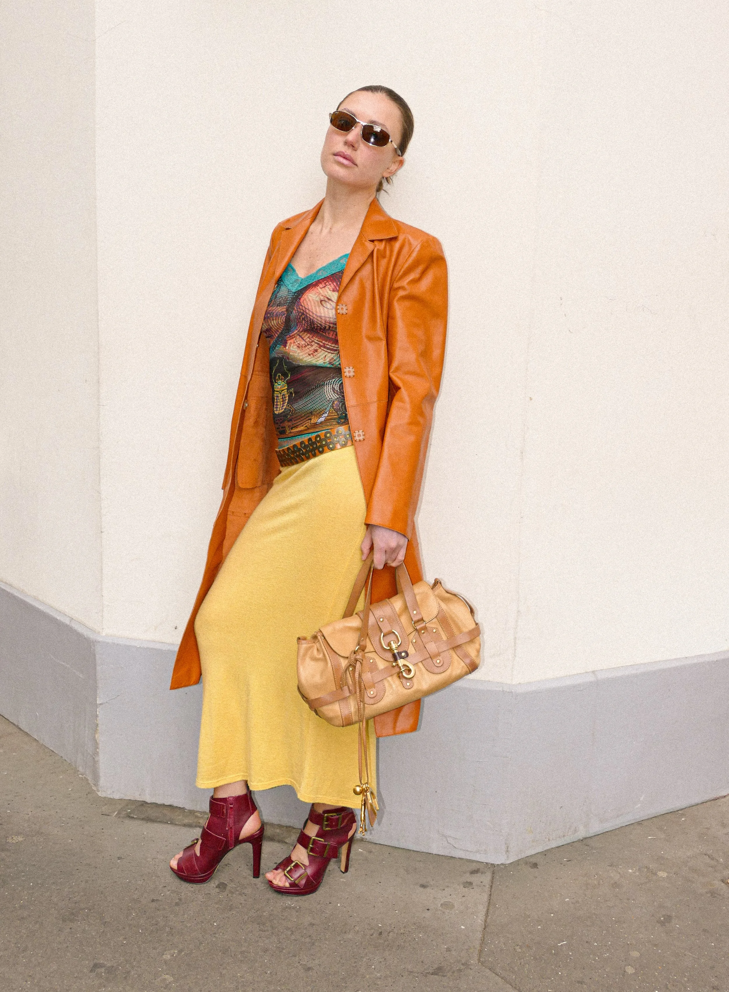Woman wearing sunglasses, an orange leather coat, a colorful top, a yellow skirt, and red high-heeled sandals standing against a plain off-white wall and holding a tan handbag.