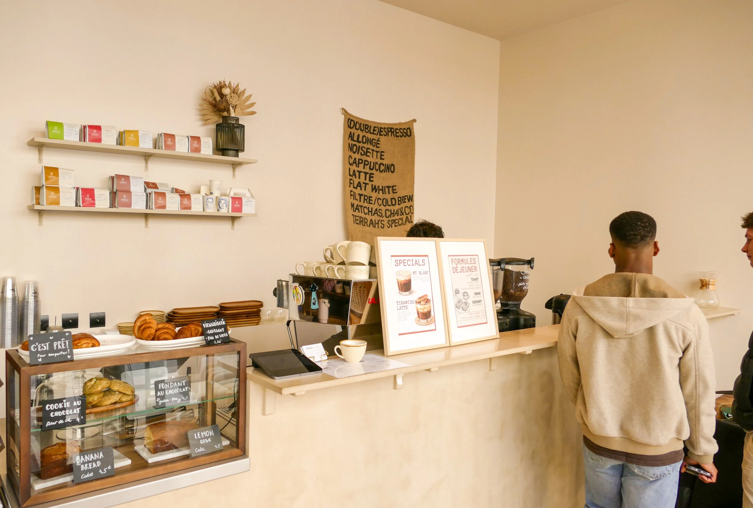 A cafe counter with a display case of baked goods, menu signs, and white shelves with coffee packages and decor. Two people are standing at the counter, one with a beige hoodie and the other with a black jacket.