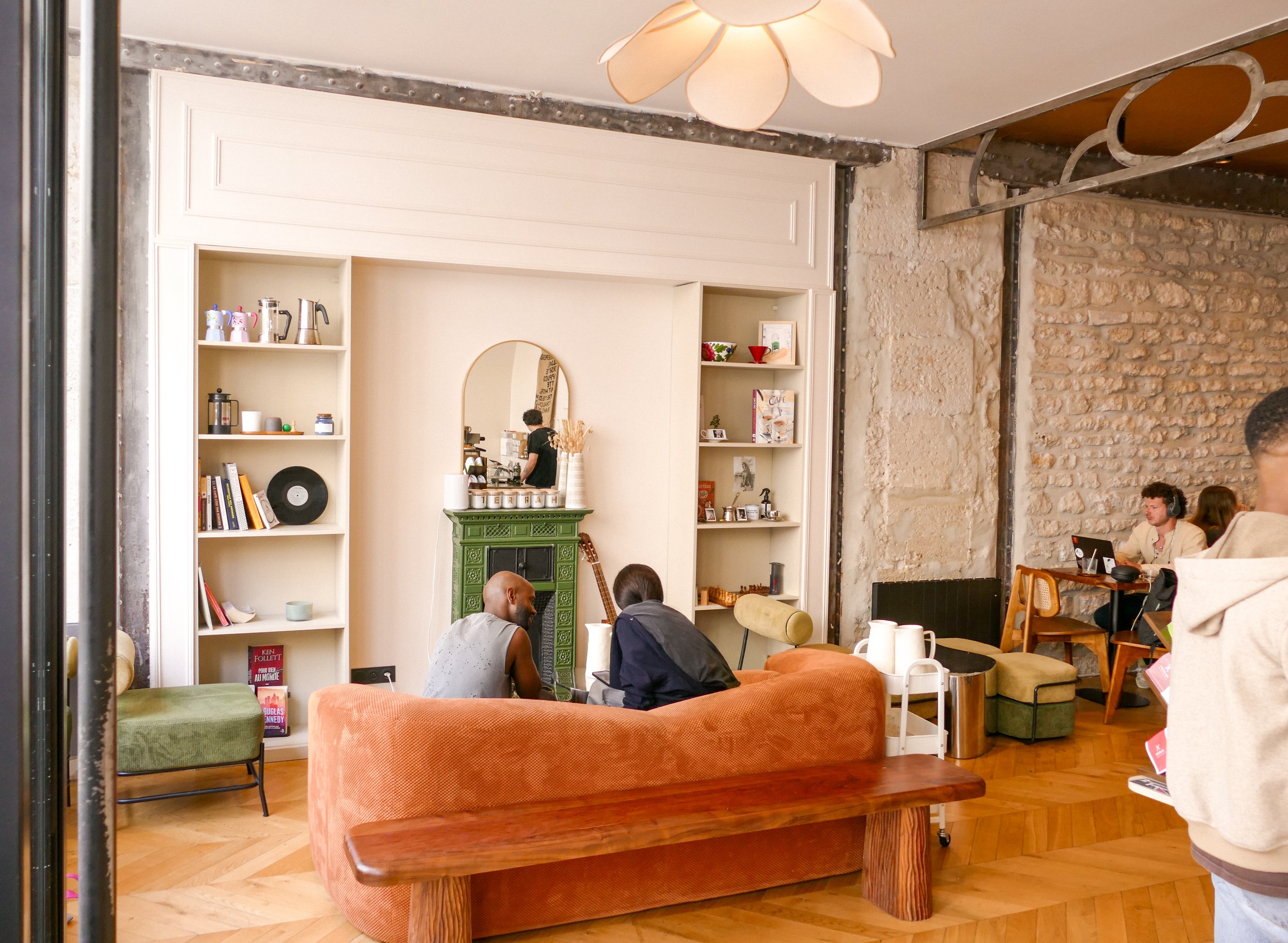 Interior of a cozy coffee shop or lounge with stone and white walls, a green fireplace, bookshelves, and people sitting on colorful cushioned chairs, one with headphones and a laptop, and a person behind the counter preparing drinks.