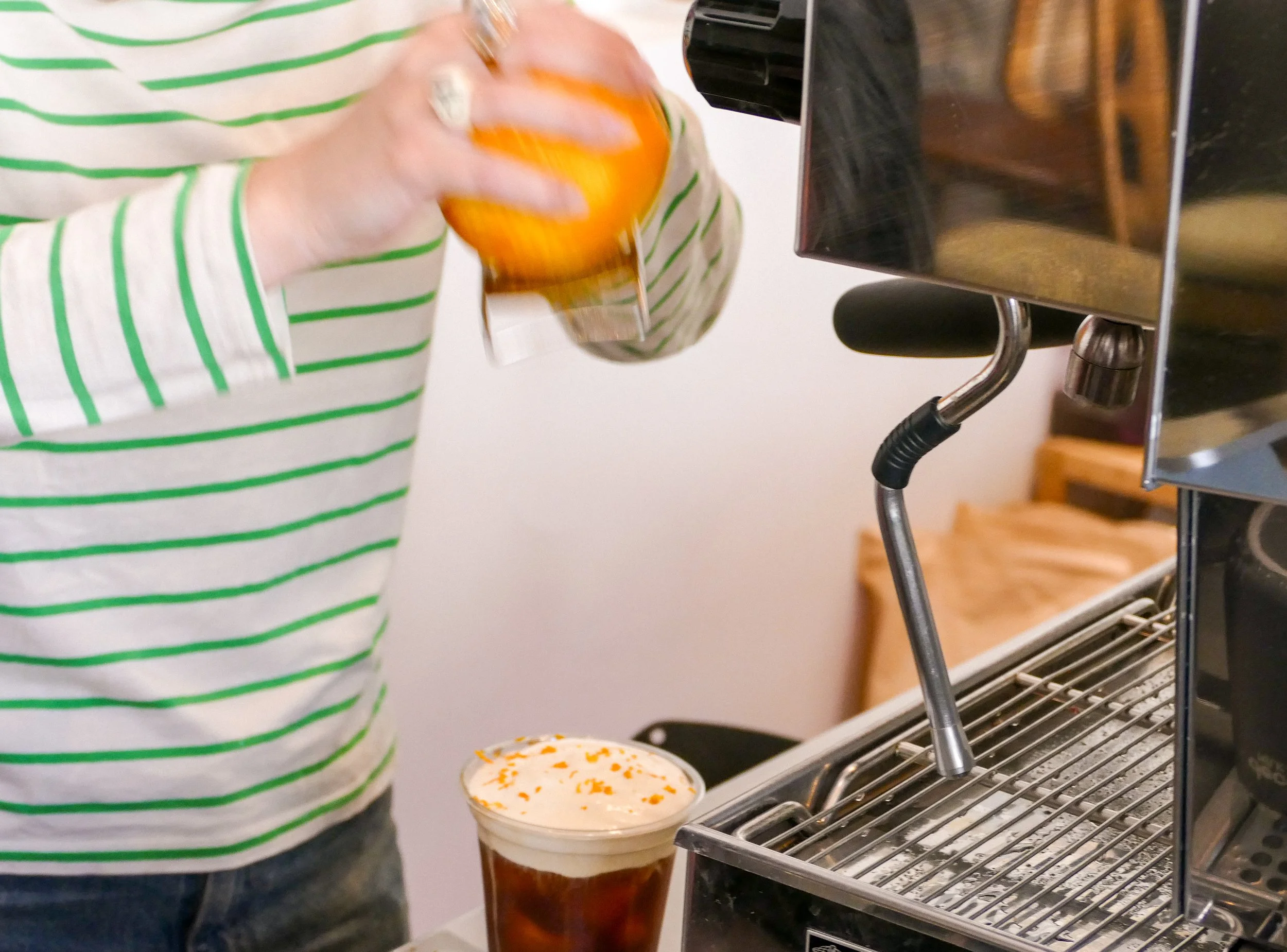 Person pouring a drink from a bottle into a glass with orange shredded topping, in front of a soda fountain machine.