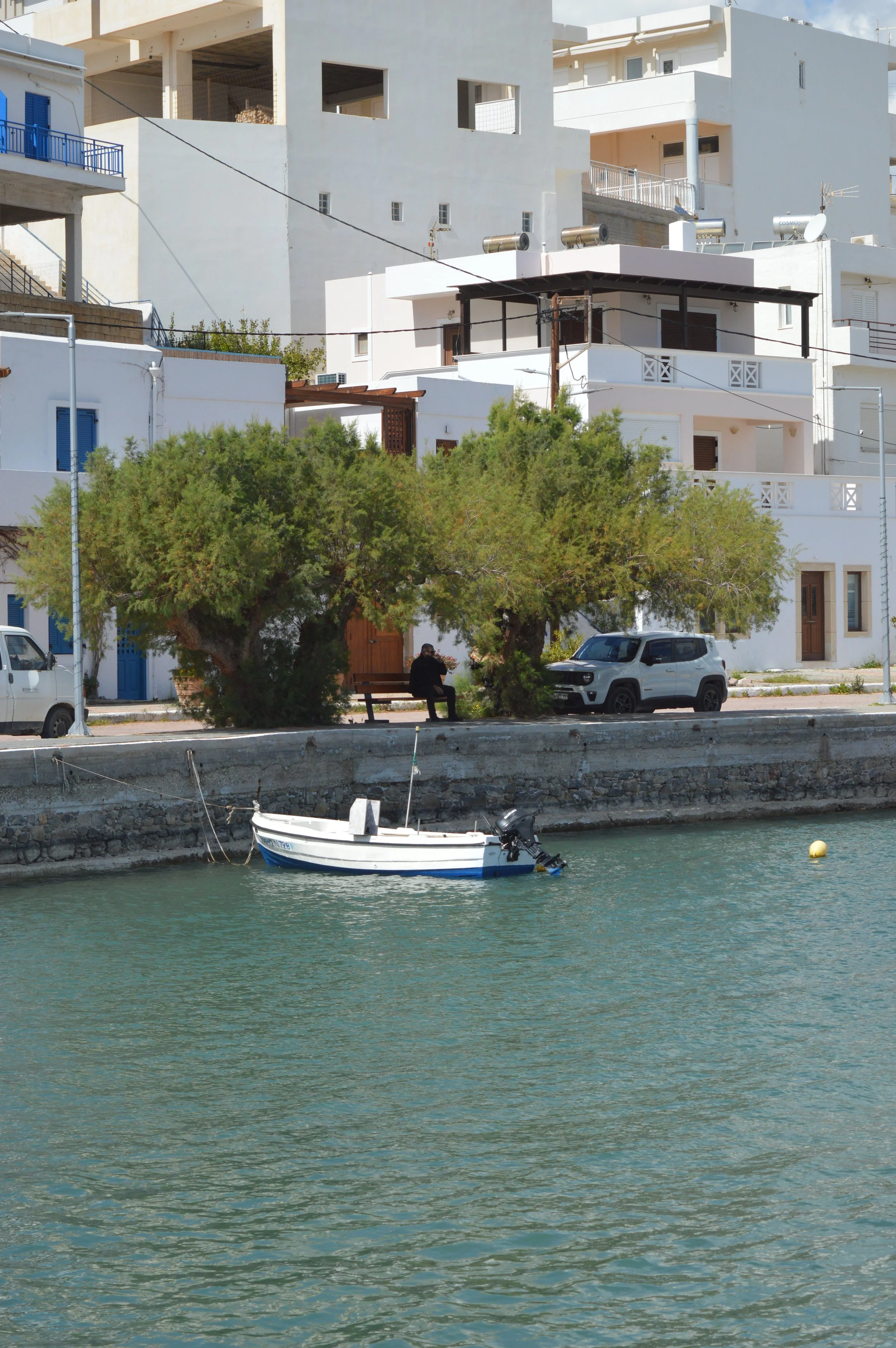 Taxi-Makry Gialos - A boat docked along a concrete shoreline, with a woman sitting on a bench underneath a tree, and white buildings in the background.