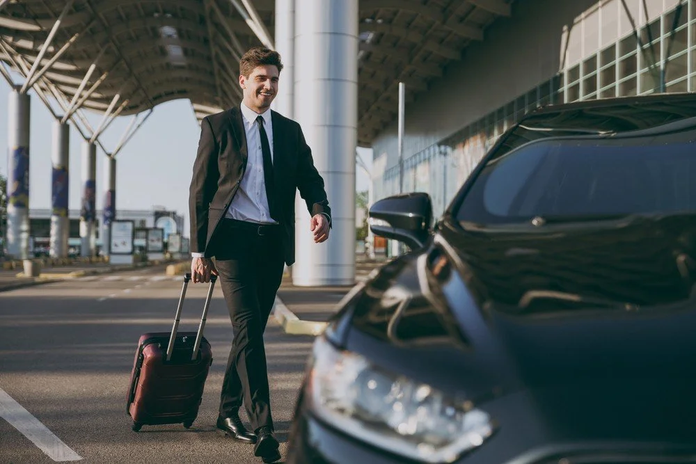 Taxi in Makry Gialos & Ierapetra - A man in a business suit walking near a black car at an airport, pulling a rolling suitcase.