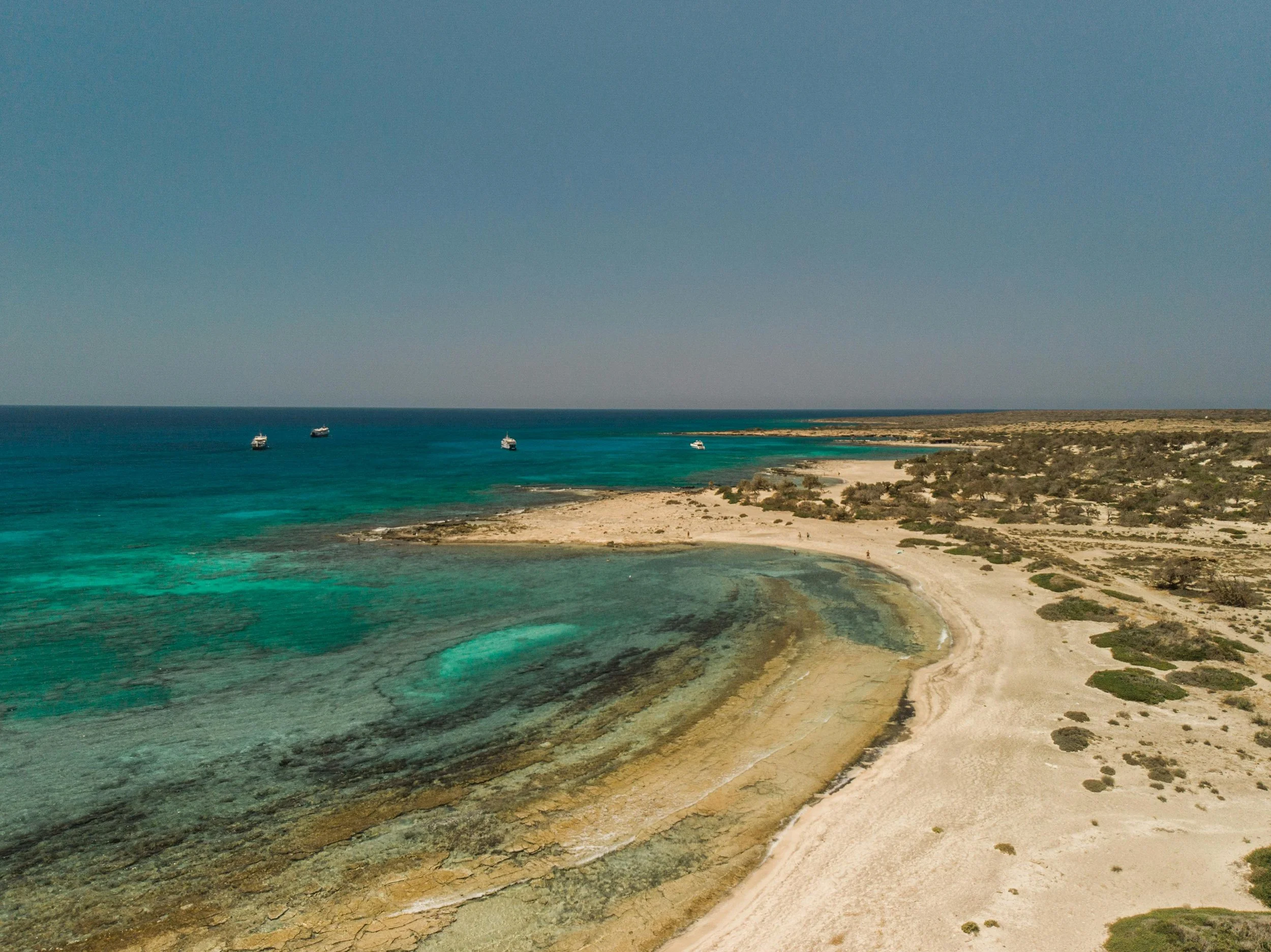 Taxi in Makry Gialos & Ierapetra - A coastal scene with clear turquoise water, a sandy beach, and a few boats floating in the water under a clear blue sky.