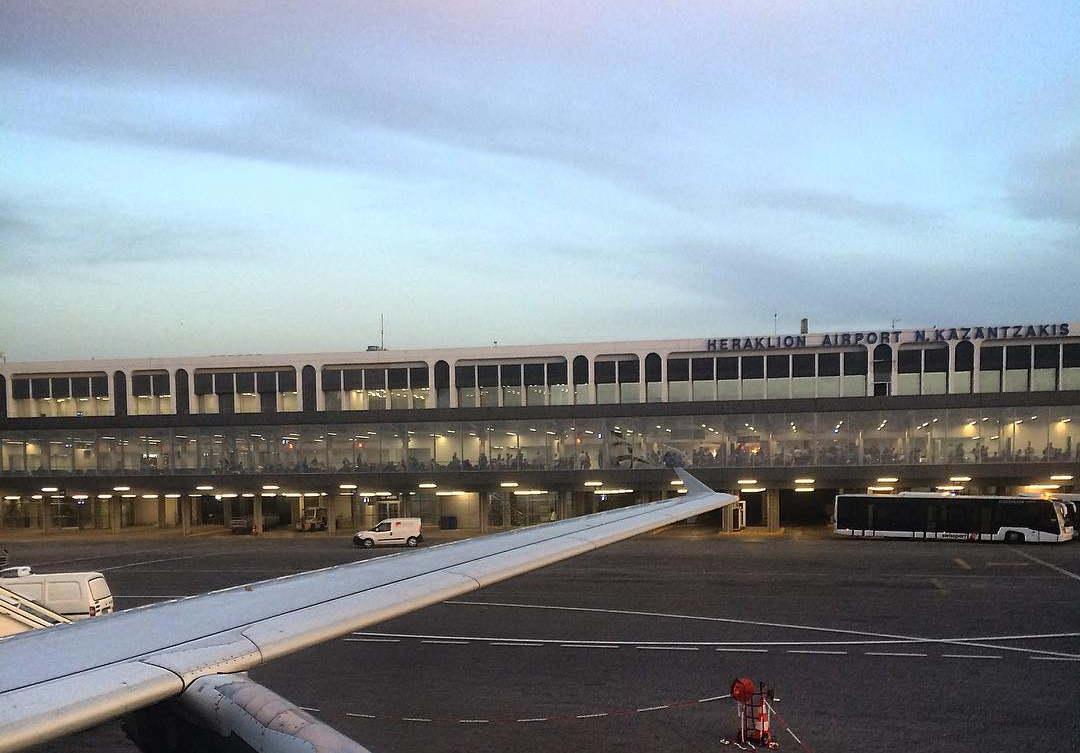 Taxi in Makry Gialos & Ierapetra - View of Heraklion Airport in Greece with airplane wing in the foreground and terminal building in the background, with several vehicles on the tarmac and people visible through the windows.