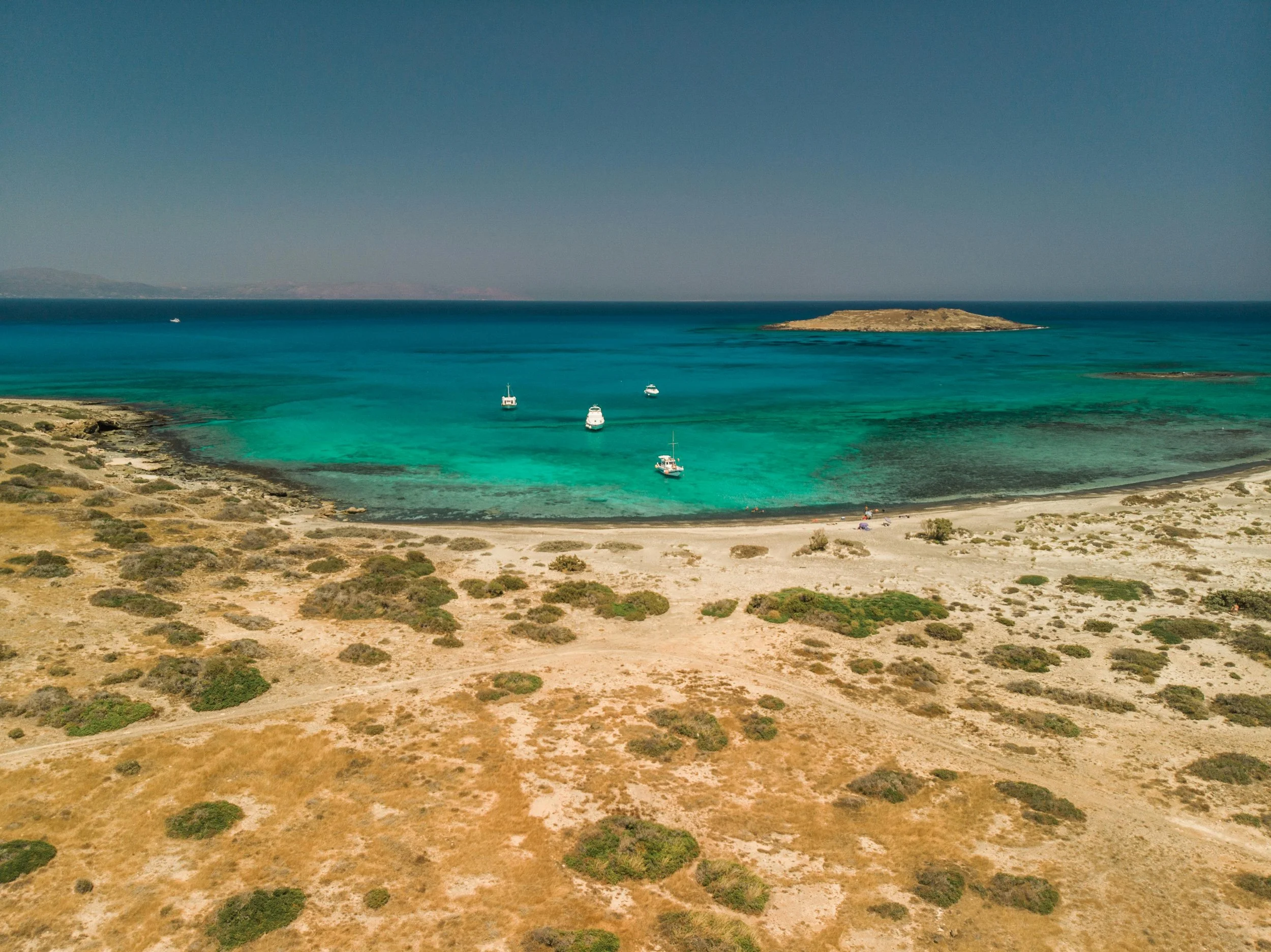 Taxi in Makry Gialos & Ierapetra - A coastal scene with turquoise water, boats anchored near the shore, and a sandy beach with sparse vegetation in the foreground. An island is visible in the distance under a clear blue sky.