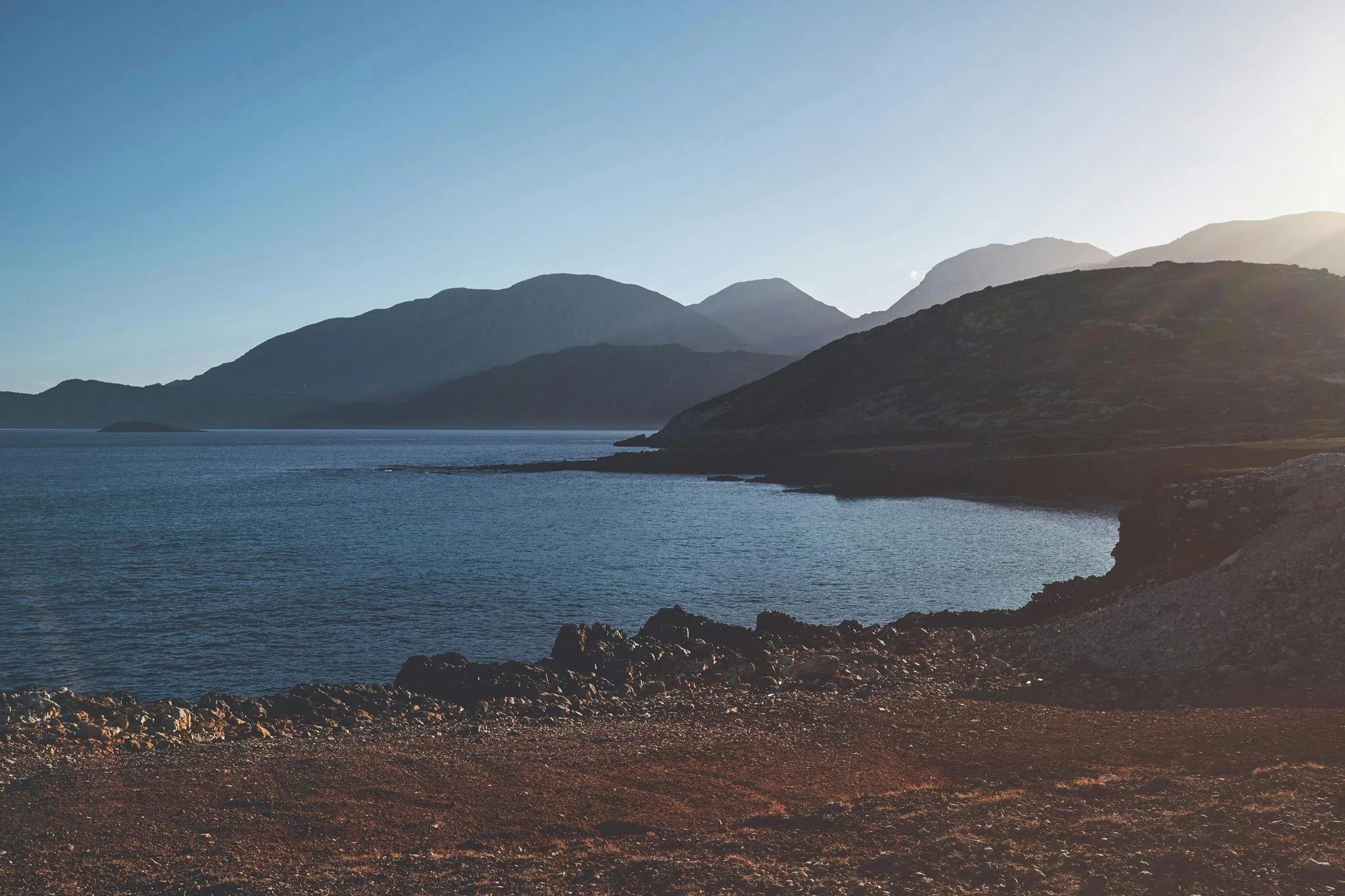 Taxi in Makry Gialos & Ierapetra - A scenic view of a lake with mountains in the background under a clear sky, viewed from a rocky shoreline.
