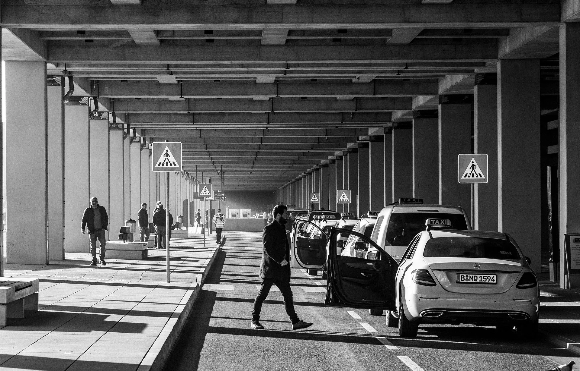 Taxi in Makry Gialos & Ierapetra - People walking and standing near parked taxis under a covered area at an outdoor transportation hub.