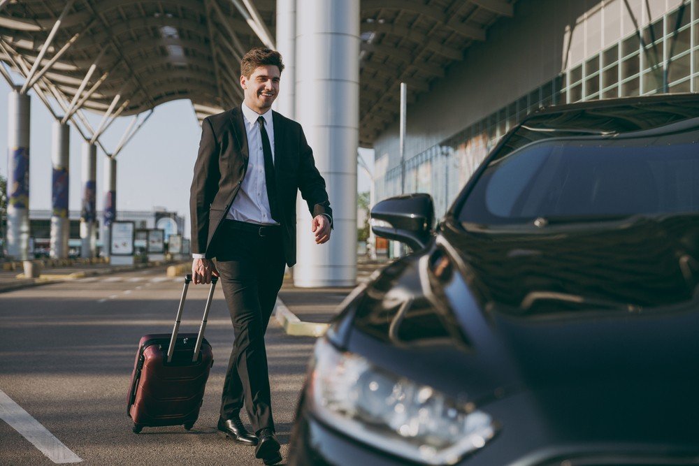 Taxi in Makry Gialos & Ierapetra - A man in a business suit walking outside an airport, pulling a rolling suitcase, with a black car in the foreground and a modern airport terminal in the background.