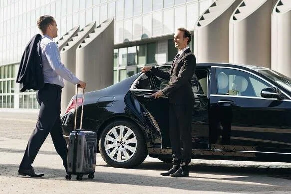 Taxi in Makry Gialos & Ierapetra - Two men talking outside an airport terminal, one with a suitcase, and the other standing by a black car.