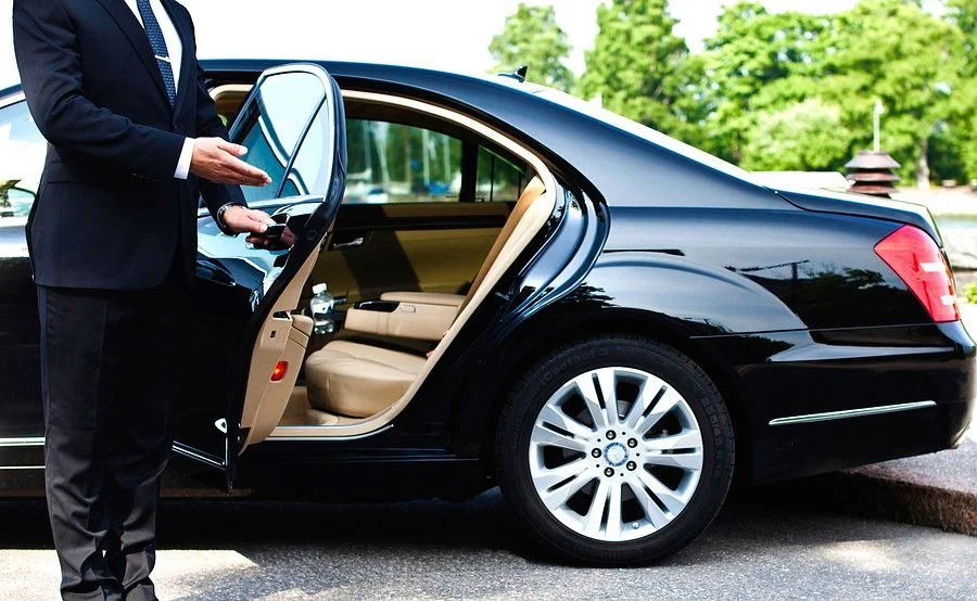 Taxi in Makry Gialos & Ierapetra - A person in a business suit standing next to a black luxury car with the rear passenger door open, revealing a beige interior.