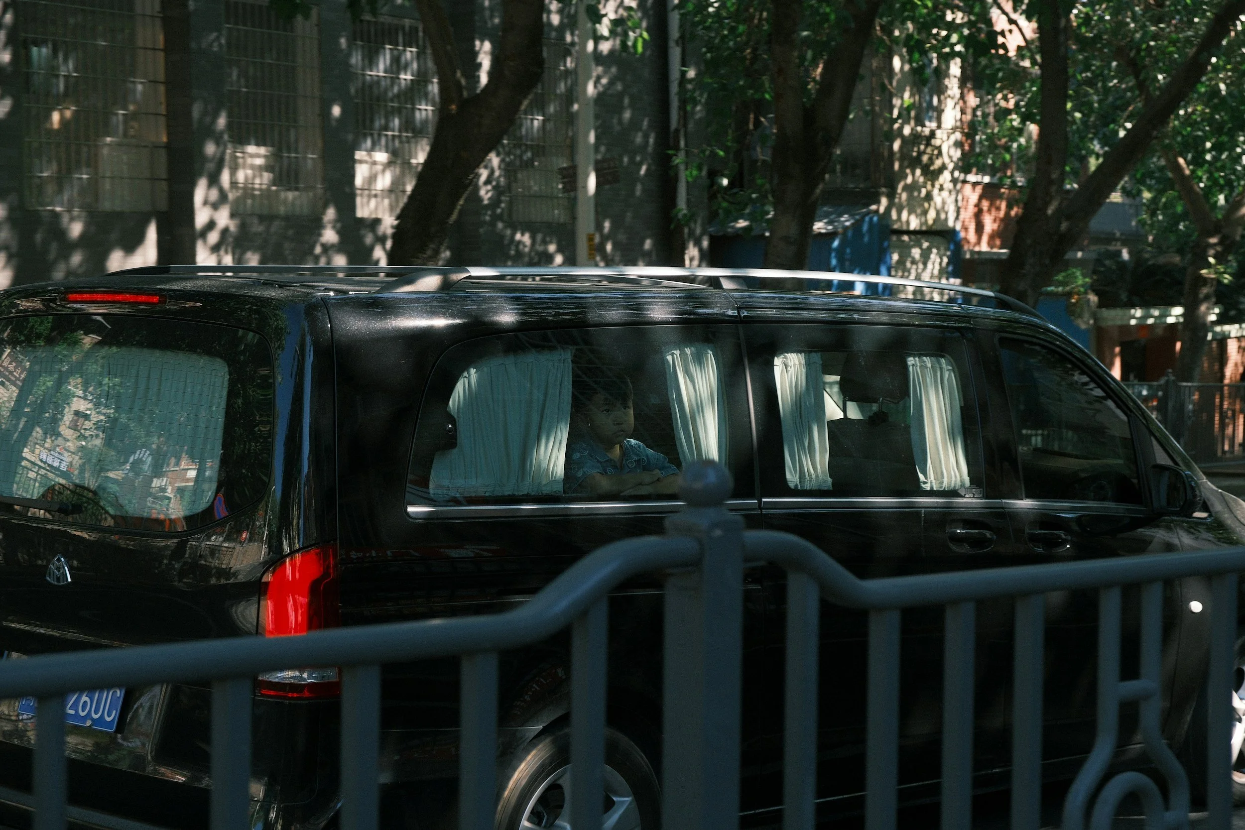 Taxi in Makry Gialos & Ierapetra - A young boy sitting inside a black SUV with white curtains on the windows. The vehicle is parked on a street with trees and a brick building in the background.