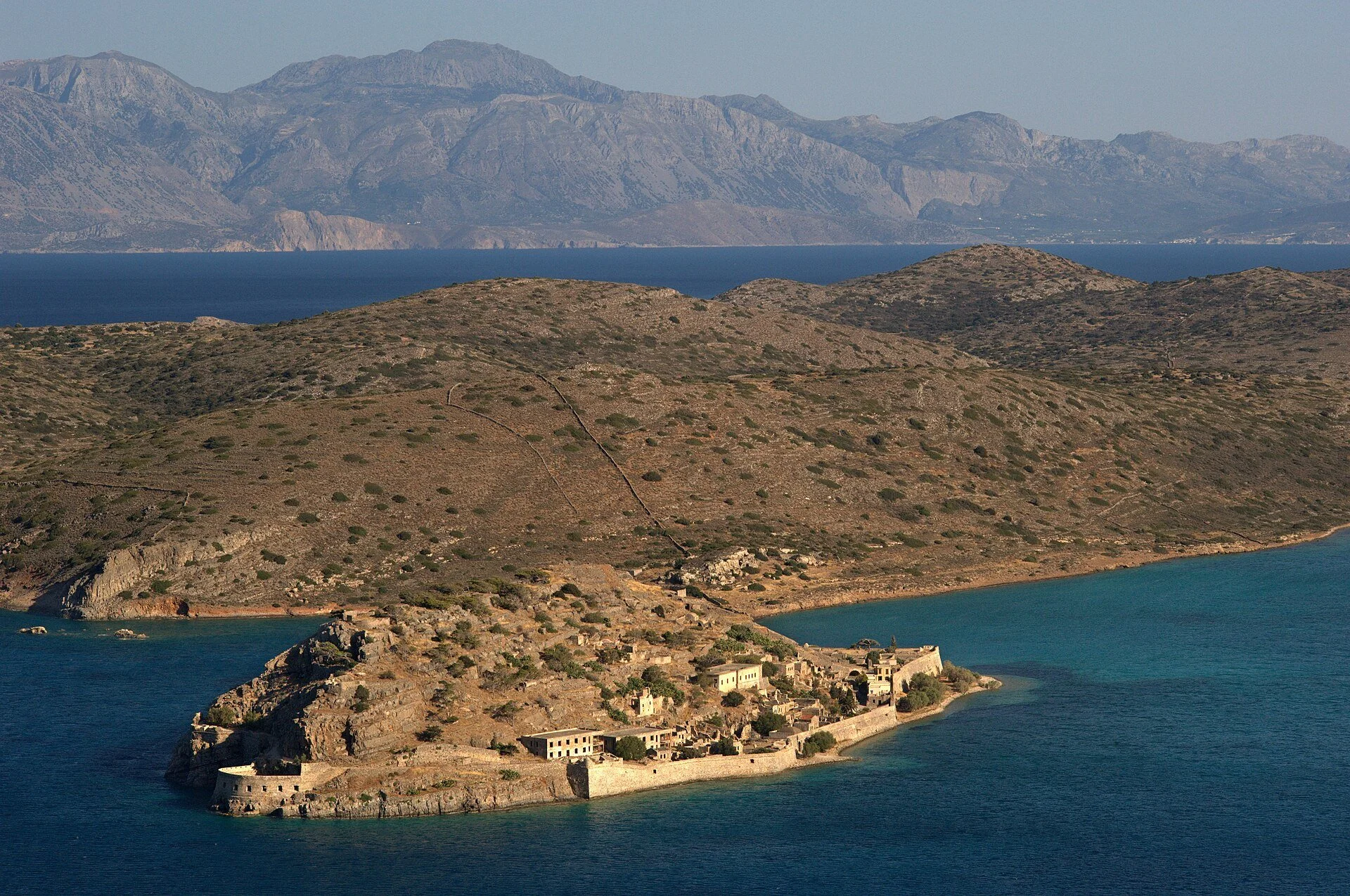 Taxi in Makry Gialos & Ierapetra - A coastal landscape with rocky hills, ancient stone buildings, and mountains in the background under a clear sky.