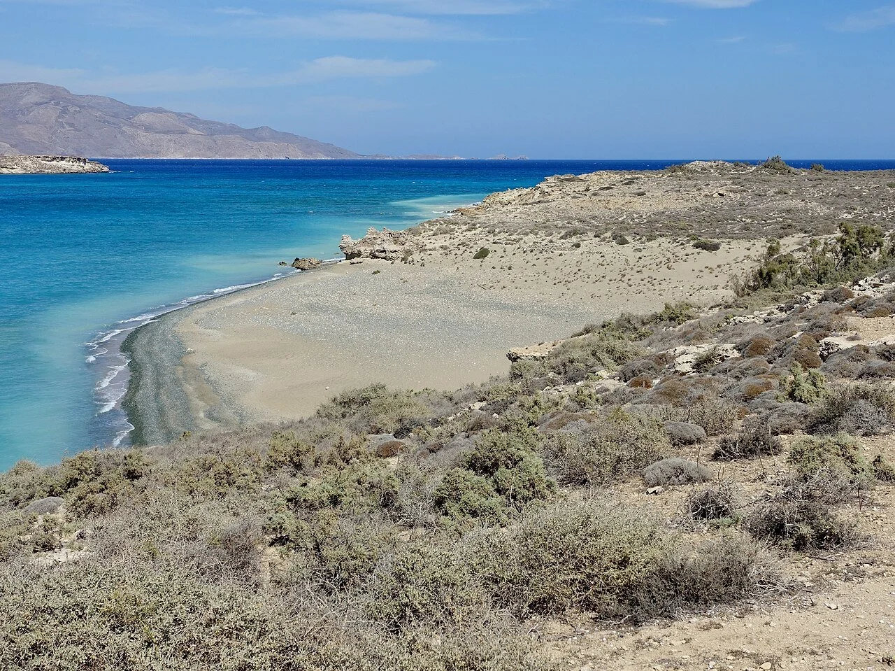 Taxi in Makry Gialos & Ierapetra - A coastal landscape featuring a sandy beach, rocky terrain, sparse desert vegetation, turquoise ocean water, and distant mountains under a partly cloudy sky.