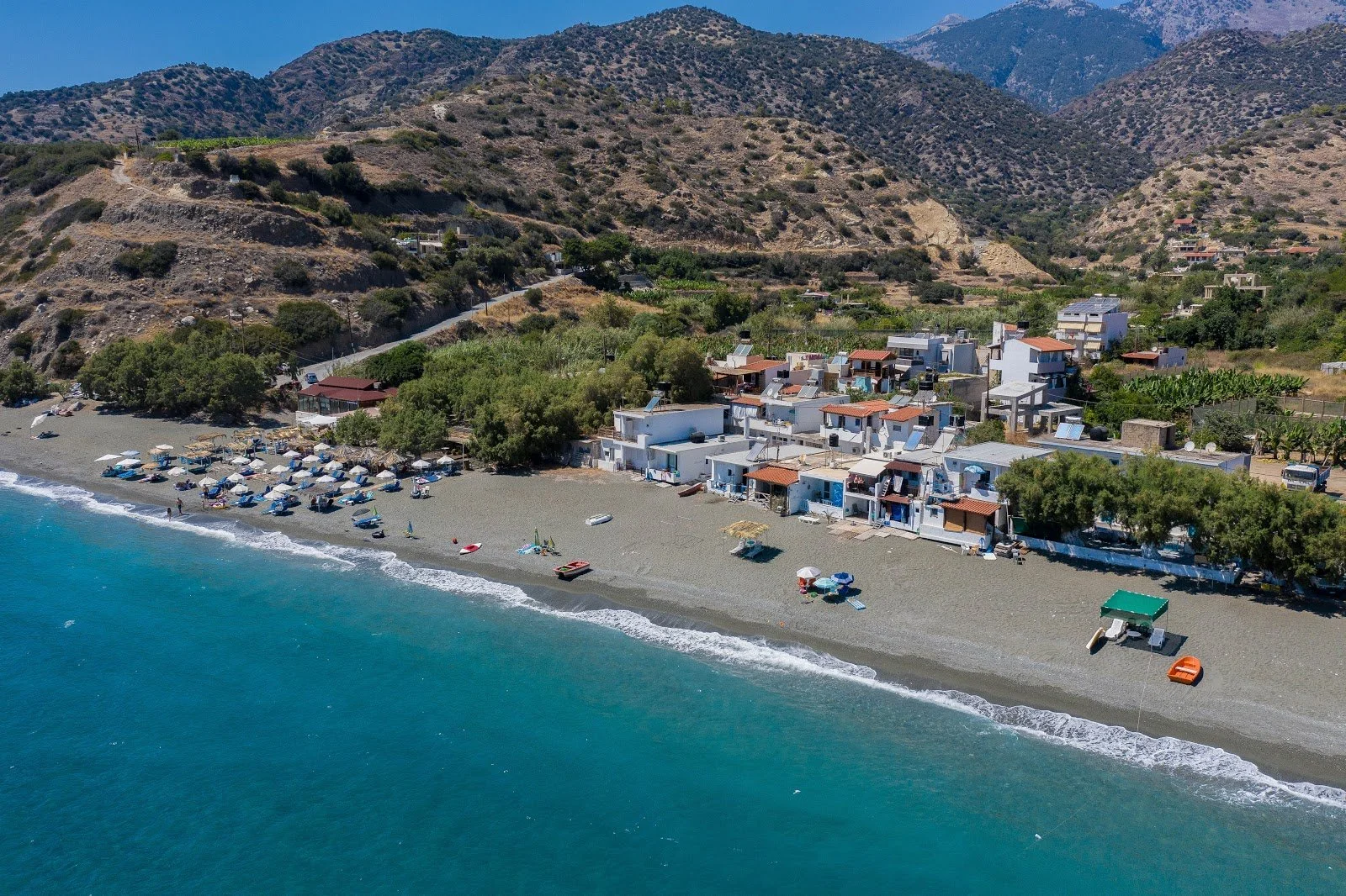 Taxi in Makry Gialos & Ierapetra - Aerial view of a small beach with umbrellas and lounge chairs, next to white houses with terracotta roofs, backed by dry hilly terrain.