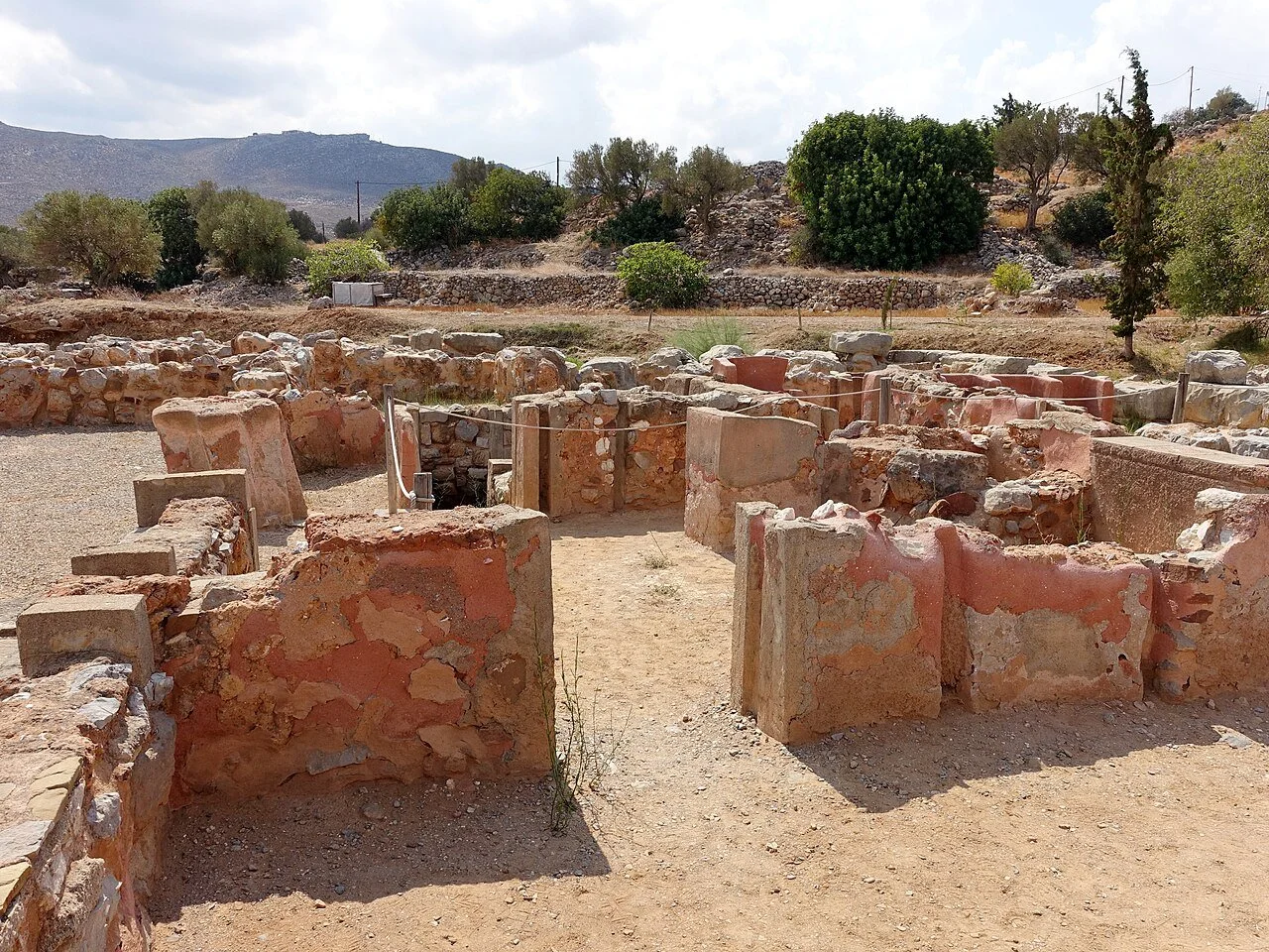 Taxi in Makry Gialos & Ierapetra - Ancient archaeological ruins with stone walls in a desert landscape, green trees and mountains in the background.