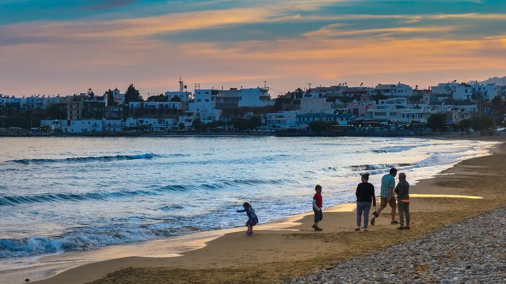 Taxi in Makry Gialos & Ierapetra - People walking along a sandy beach during sunset, with waves gently rolling in and a town with white buildings in the background.