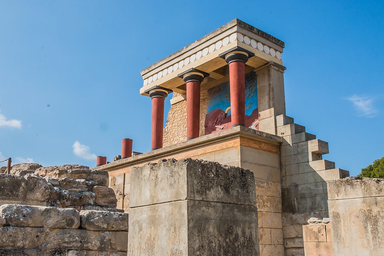 Taxi in Makry Gialos & Ierapetra - Ancient ruins with a small temple featuring red columns and a mural, set against a blue sky.