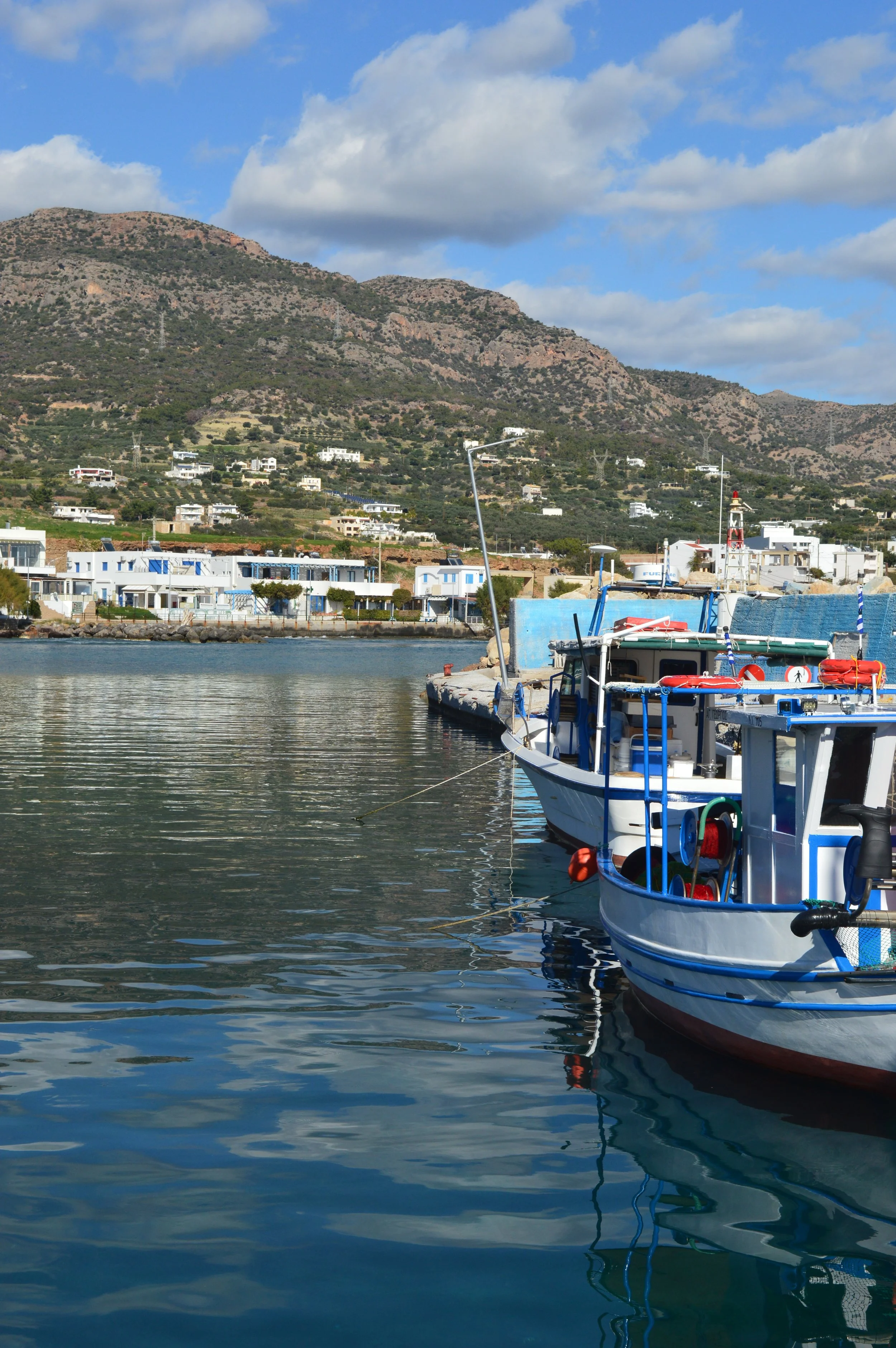 Taxi-Makry Gialos - Boats docked in a harbor with calm water, white modern buildings along the shoreline, hills with sparse vegetation and scattered houses in the background, under a partly cloudy sky.