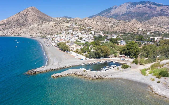 Taxi in Makry Gialos & Ierapetra - Aerial view of a coastal town with a beach, boats, and a harbor, surrounded by mountains under a clear blue sky.