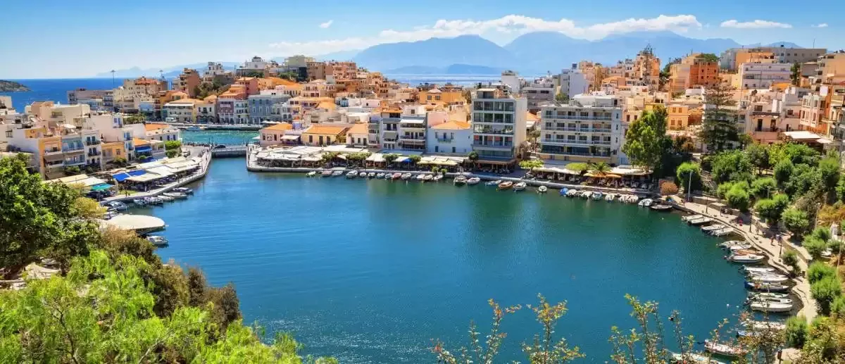 Taxi in Makry Gialos & Ierapetra - A scenic harbor with calm blue waters surrounded by colorful buildings, docked boats, and lush greenery in a Mediterranean town with mountains in the background.