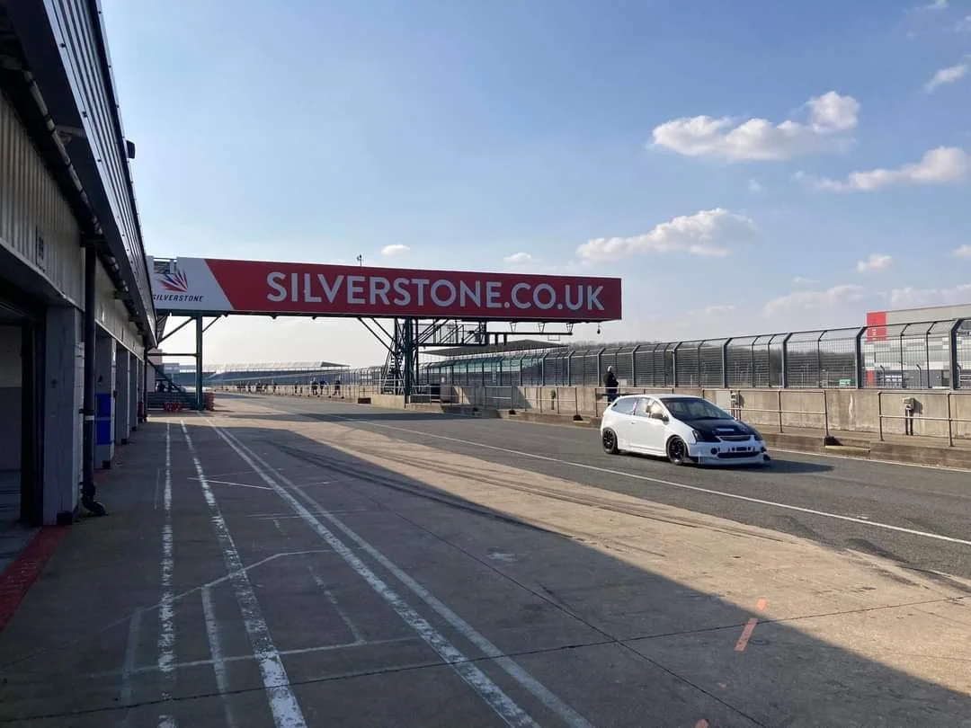 Empty motorsport pit lane at Silverstone race track with a silver race car parked, blue sky with clouds, and large red banner advertising silverstone.co.uk.