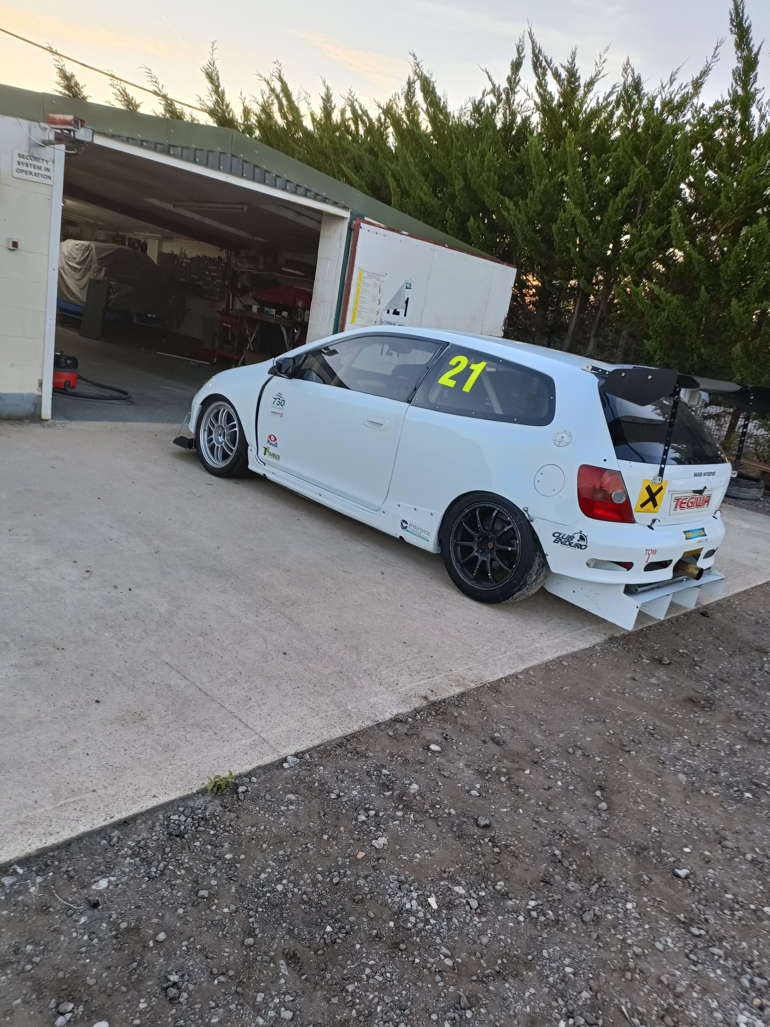 A white race car with the number 21 on its side, parked on a gravel area outside a garage with a tarp-covered vehicle and tools inside.