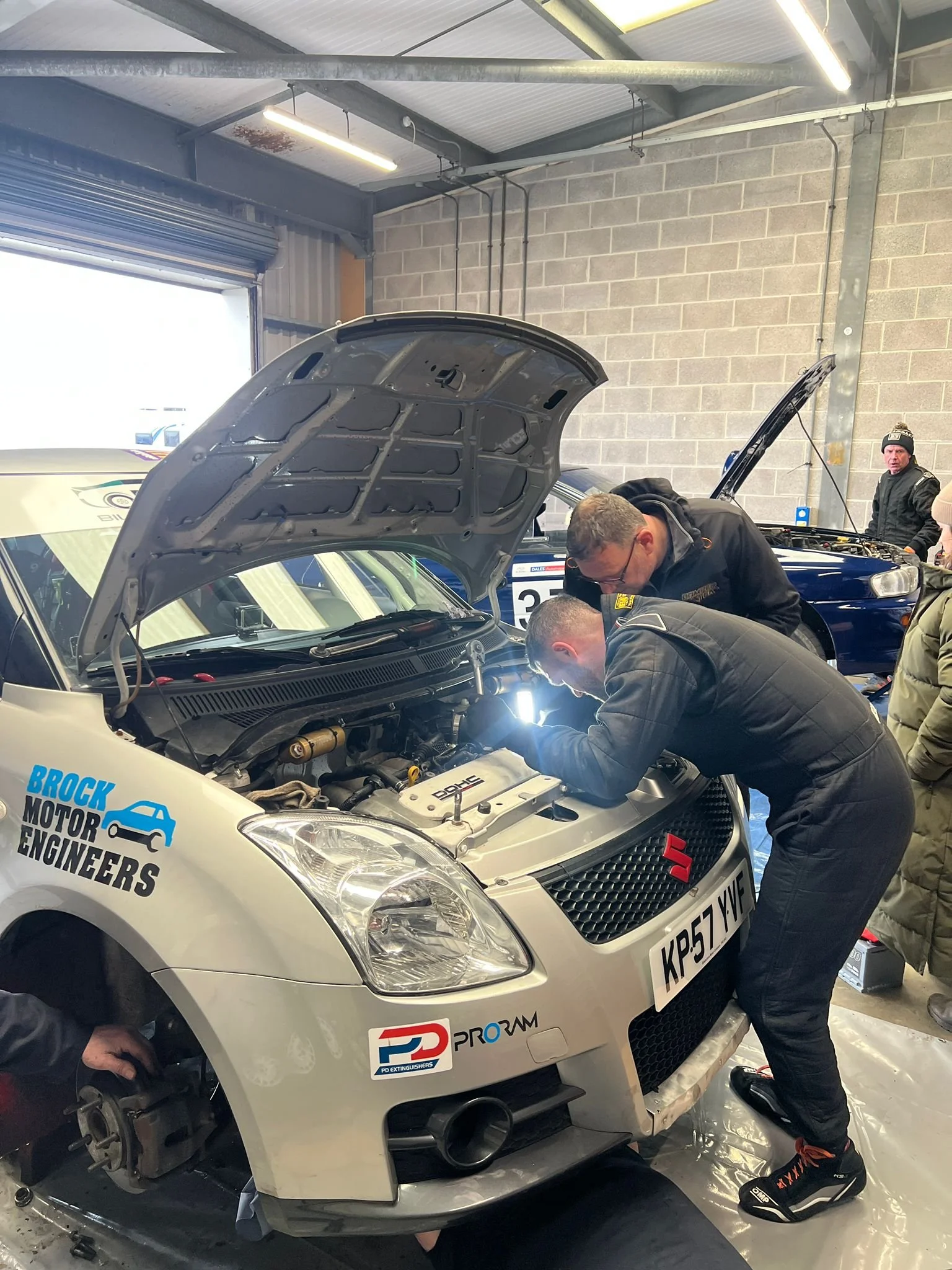 Several people working on a white race car with its hood open inside a garage. The car has various stickers, including one that says 'Broock Motor Engineers' and 'PD PROGRAMES'. One person is leaning over the engine, and another person is standing in the background near another blue car.