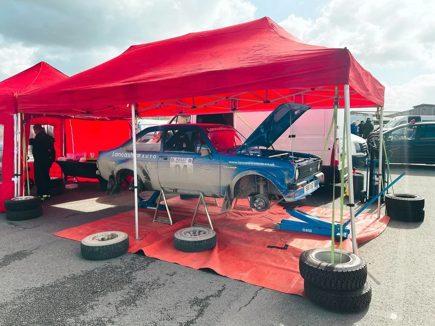Rally car on a red tarp under a red canopy, with wheels removed and the hood open, at a pit stop or service area.