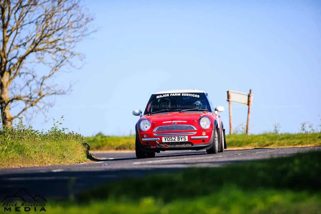 A red Mini Cooper race car on a rural road with a grassy landscape and clear blue sky.