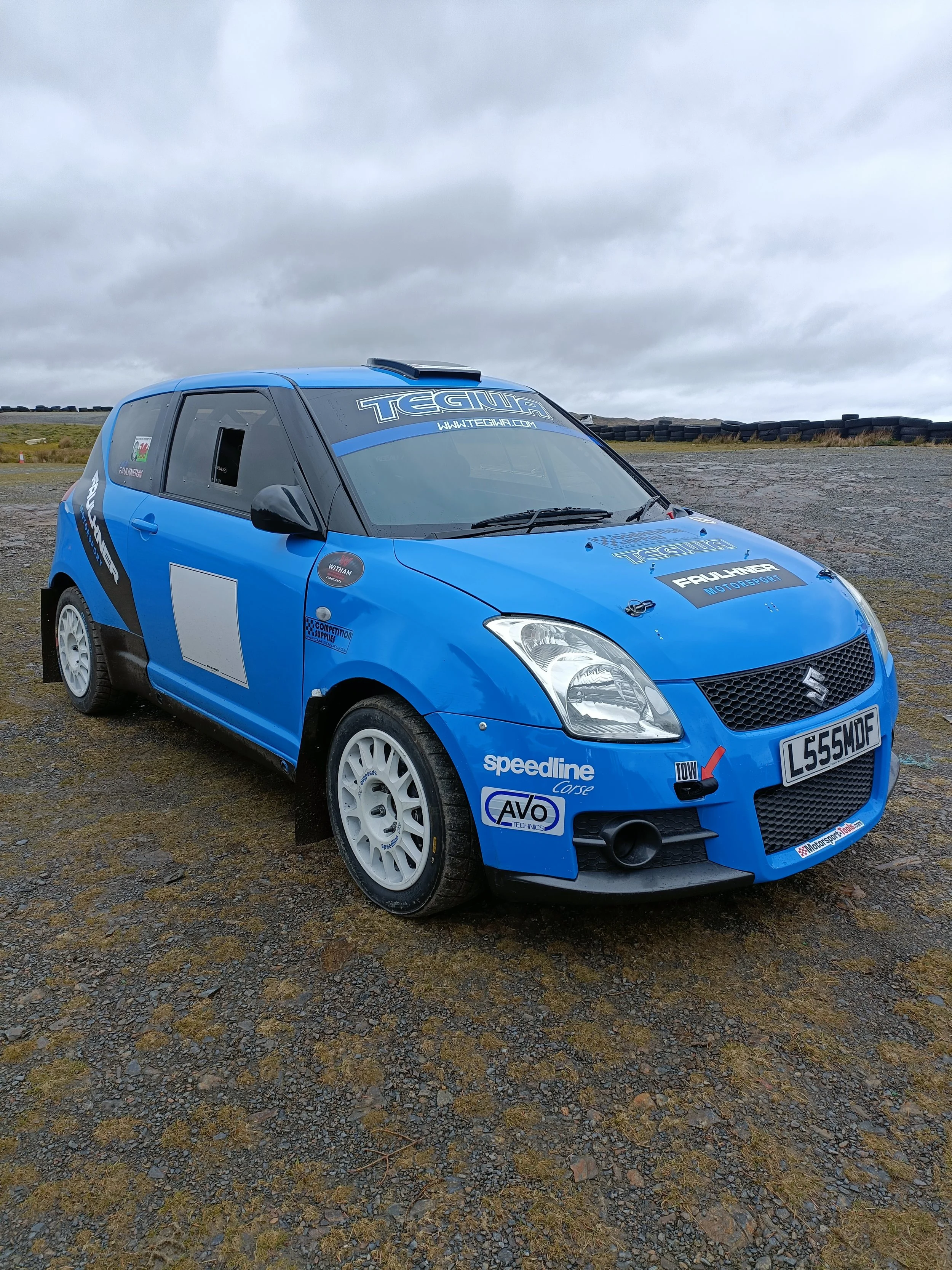 A blue racing car with sponsor stickers and a license plate, parked on a gravel area outdoors under a cloudy sky.