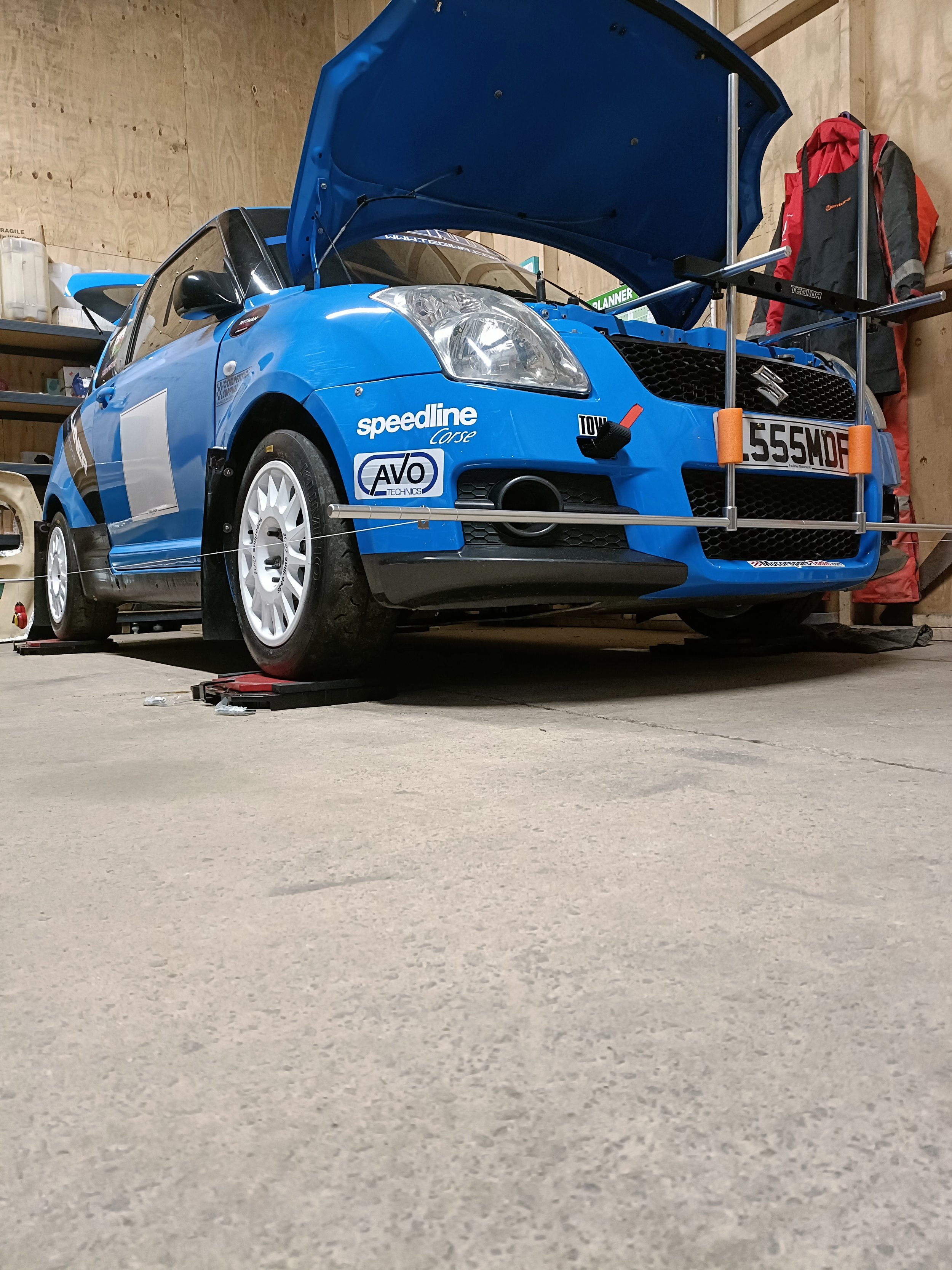 A blue racing car with a hood open, supported by a metal prop, inside a garage. The car has various sponsor logos and a license plate '555MDP'.