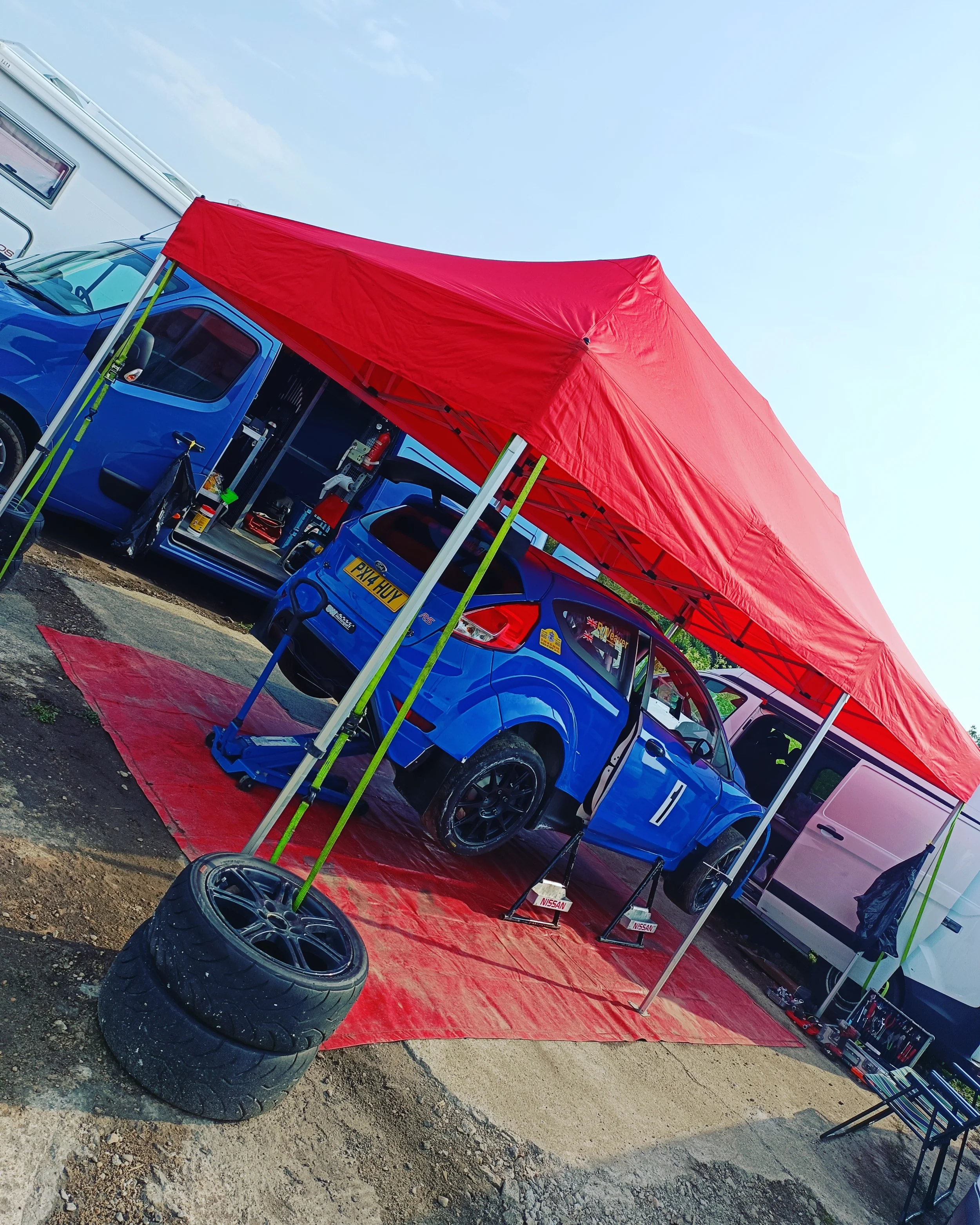A blue rally car parked at a racing event under a red canopy, with spare tires in front and tools on a table nearby.