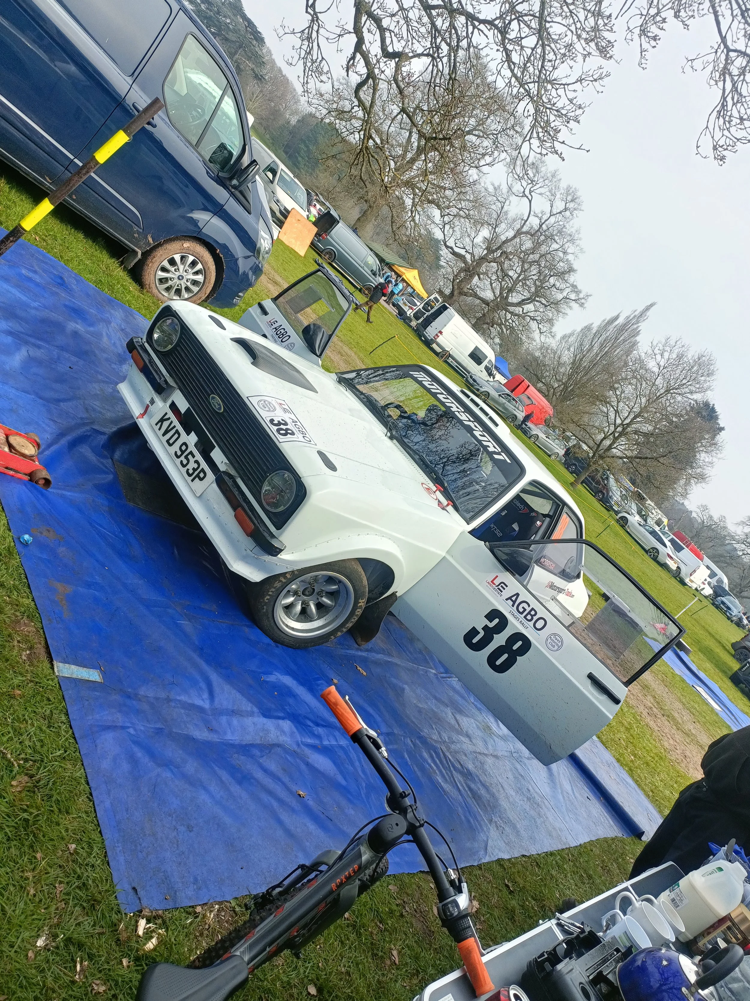 A white race car with the number 38 and various sponsor decals on the side, parked on a blue tarp in an outdoor area with multiple other vehicles, trees, and a tent in the background.