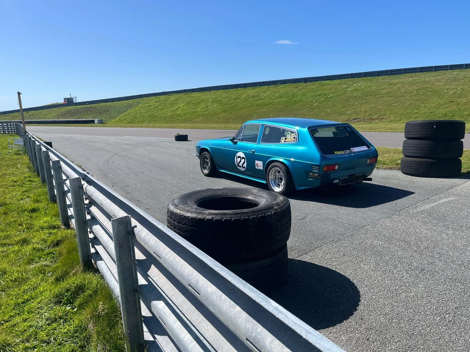 A vintage blue race car with the number 122 on the door, parked on a racetrack with tires stacked nearby and a guardrail on the side, under a bright blue sky.