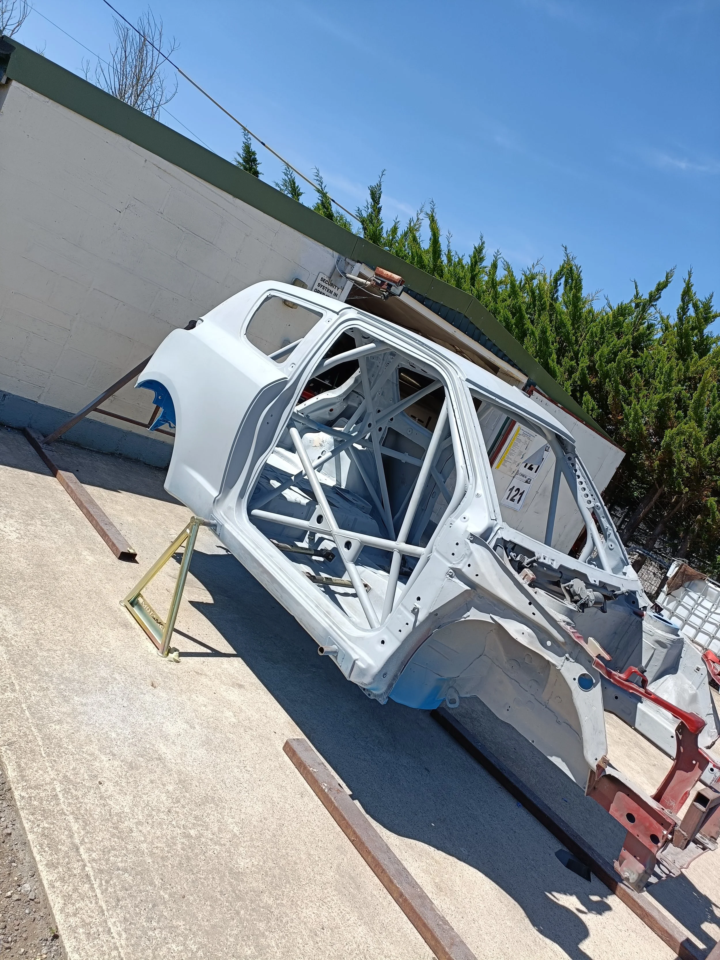 A white car chassis on a trailer, with an open door and visible roll cage, parked outdoors next to a wall and trees.