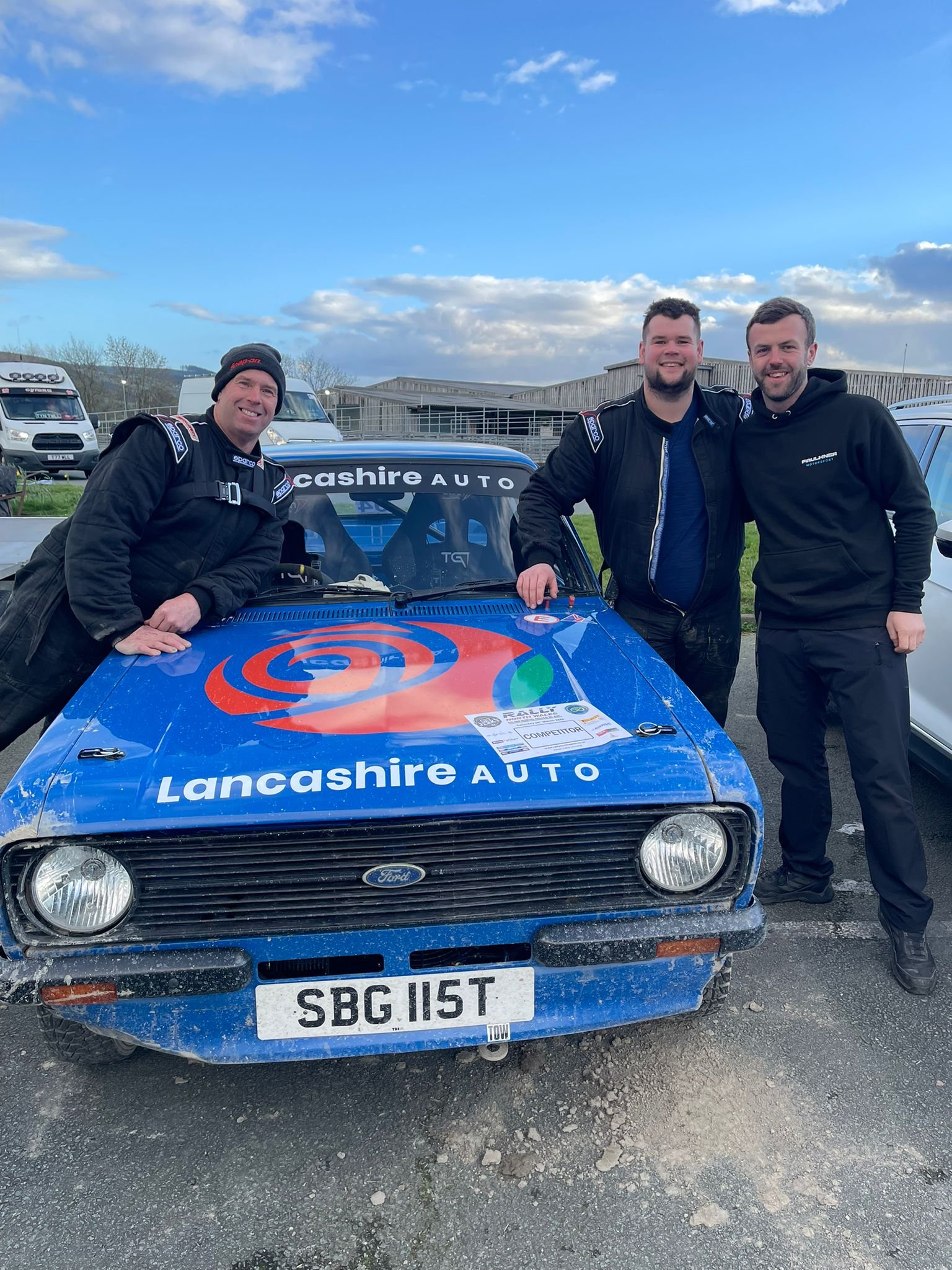 Three men are standing next to a blue rally car with 'Lancashire AUTO' written on the windshield. Two of the men are wearing black racing suits, and one is wearing a black hoodie. All three are smiling. The scene is outdoors, with a partly cloudy sky and some vans and buildings in the background.