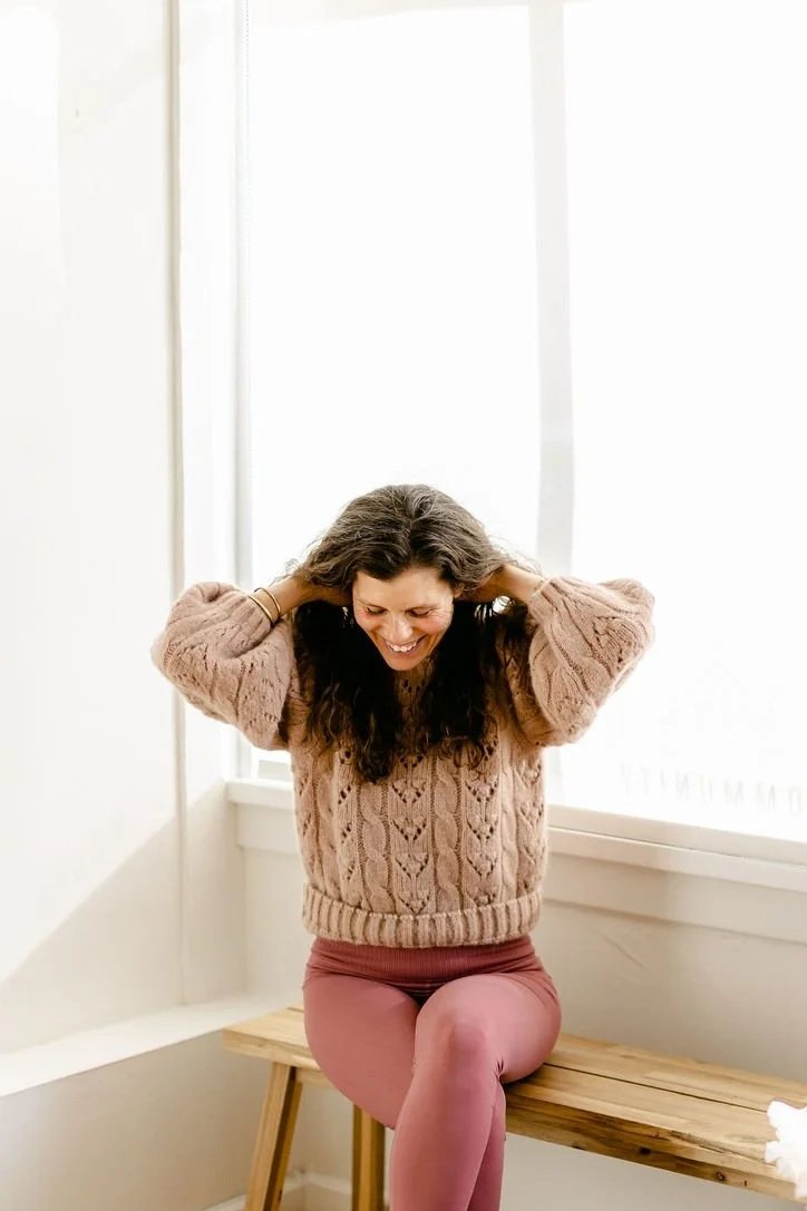 A woman with brown hair sitting on a wooden bench, smiling, with her hands behind her head in a bright room with a large window.