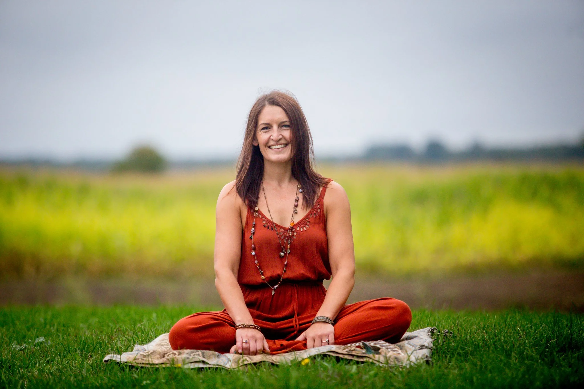 A woman with brown hair and a red dress sitting cross-legged on a blanket in a grassy field, smiling at the camera with a blurred green and gray background.