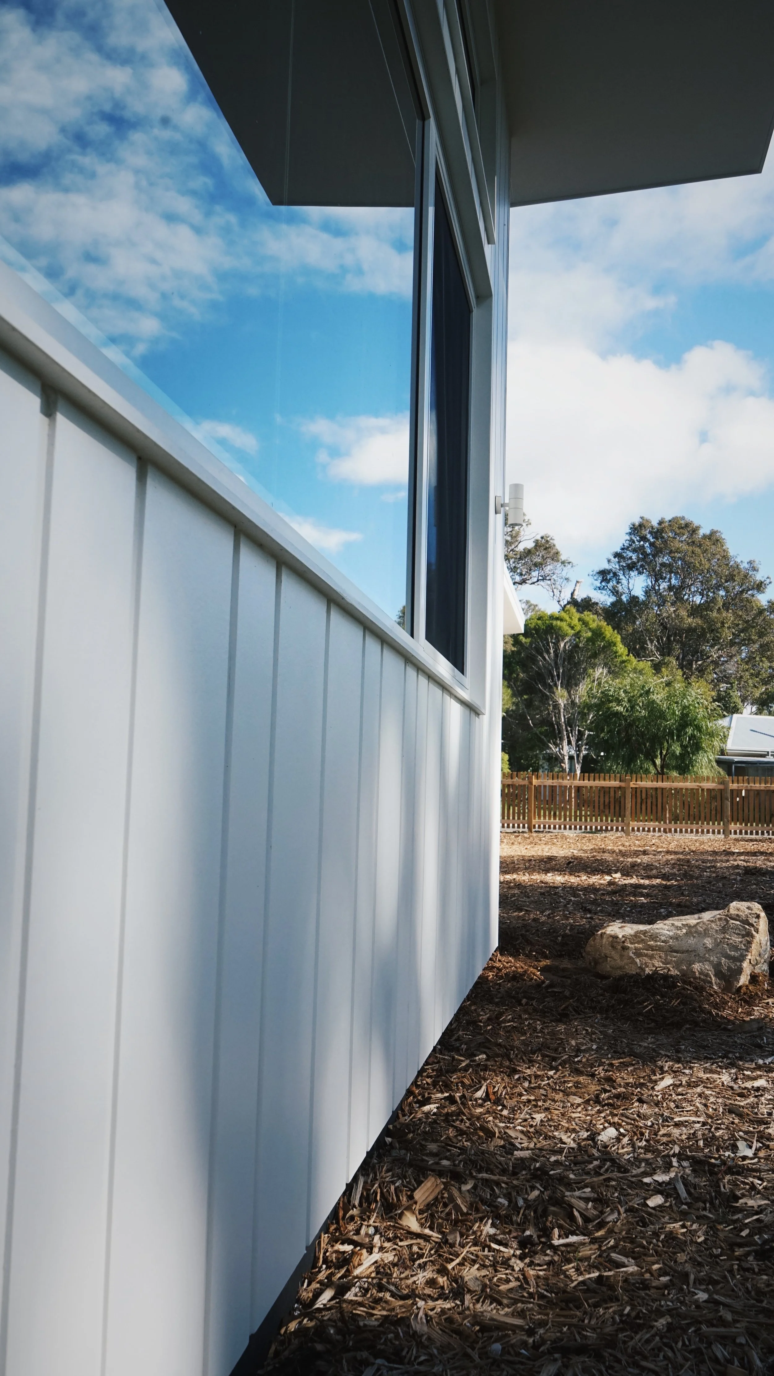Exterior view of a modern house with reflective windows, white vertical siding, surrounded by mulch and a wooden fence, with trees and a blue sky with clouds in the background.