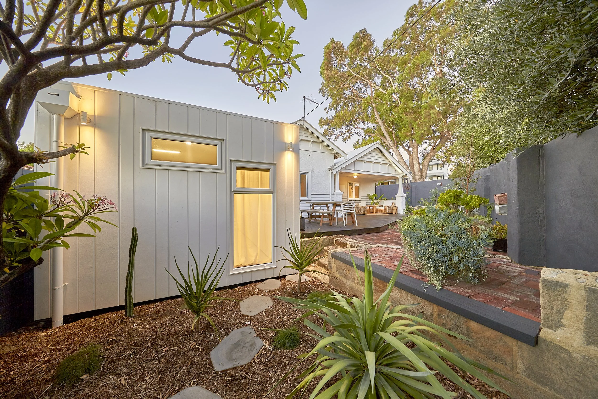 A modern white house with a backyard garden, wooden deck, outdoor seating, surrounded by lush green plants and trees, with a gray wall and clear sky in the background.
