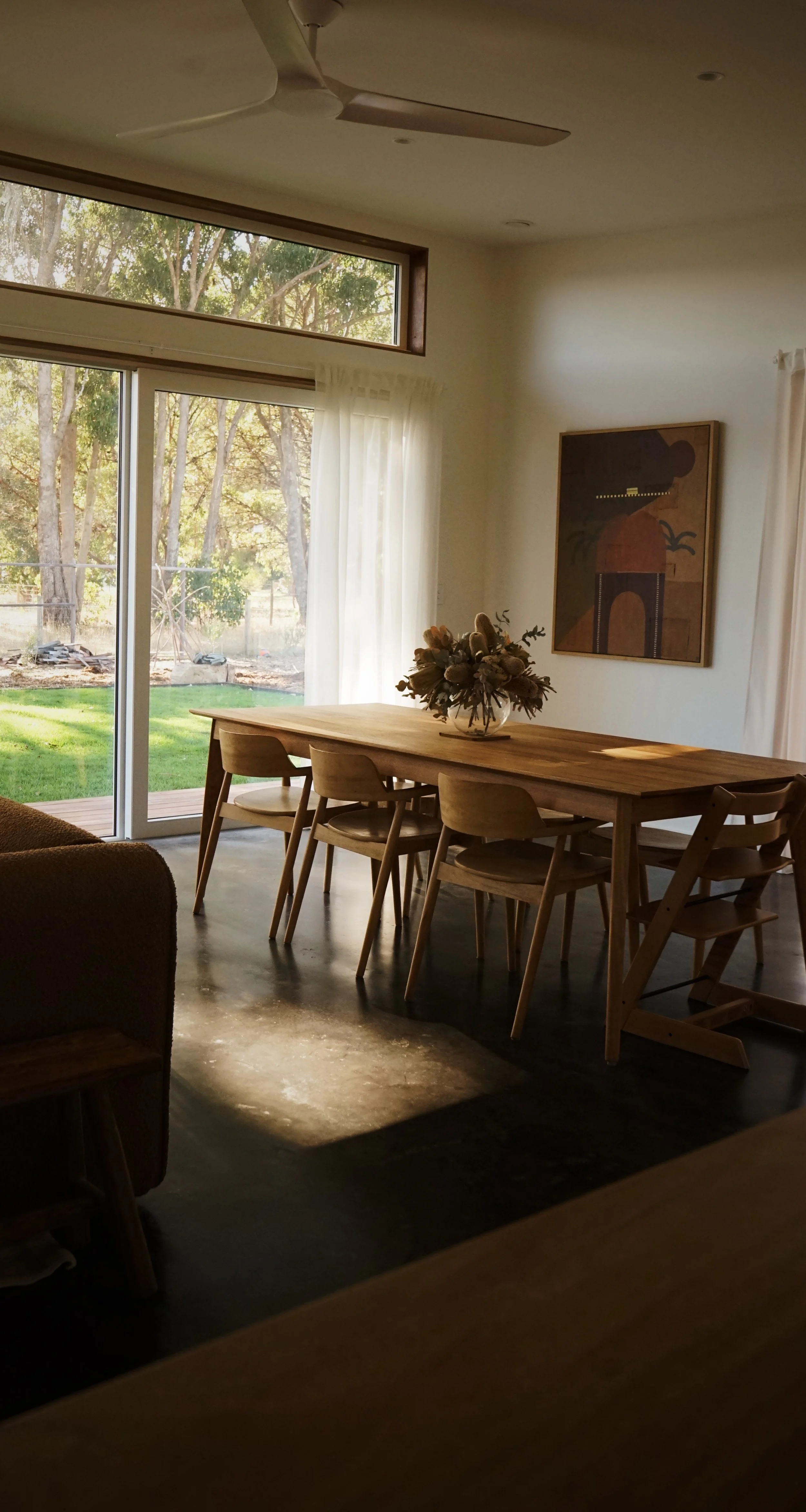 A dining room with a large wooden table and eight chairs, a vase of flowers in the center, large windows with white curtains, and a view of a backyard with trees and grass.