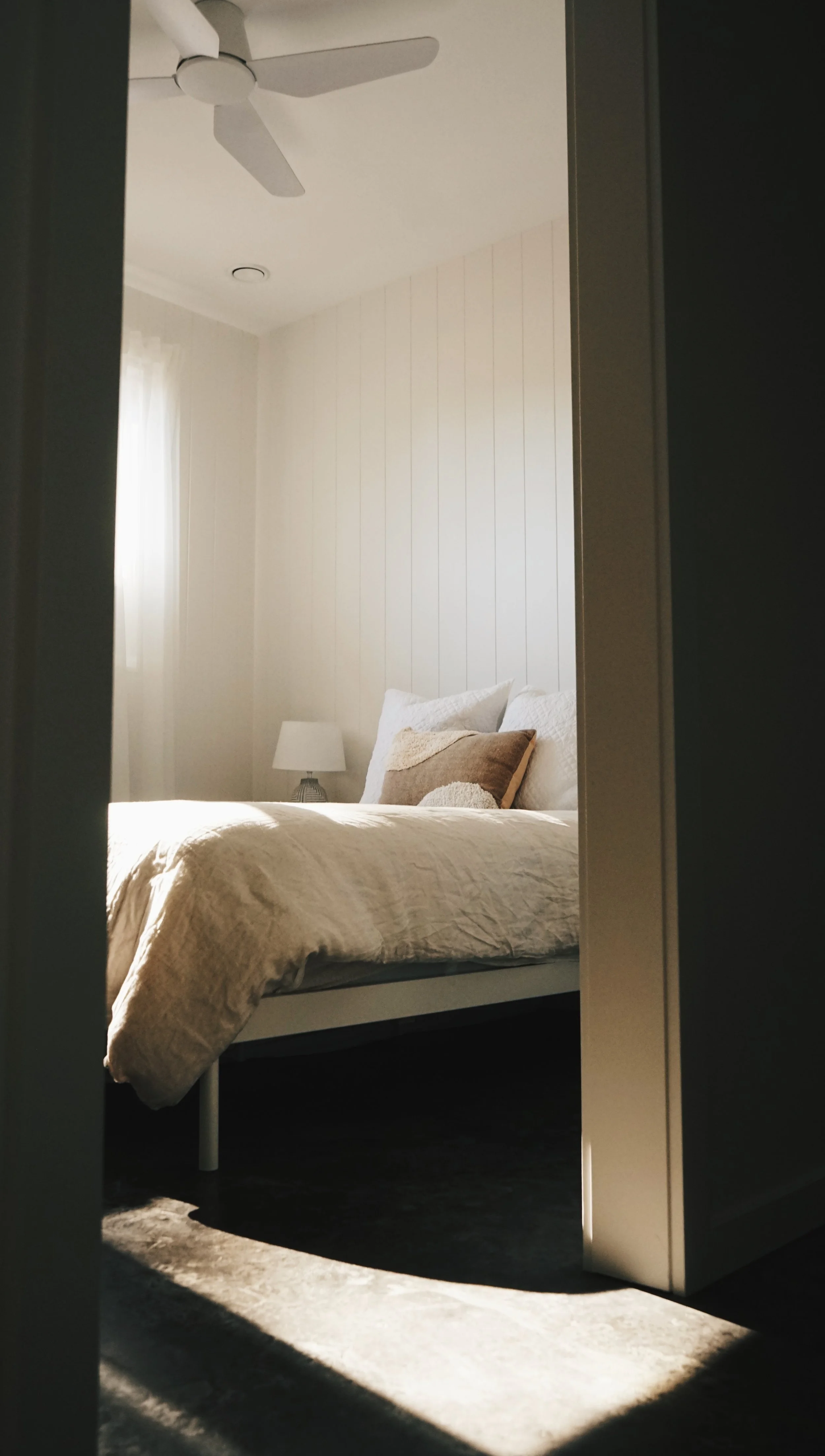 A bedroom viewed through a doorway, with a bed made with beige bedding and pillows, a nightstand with a lamp, and a window with sheer curtains in the background. Sunlight casts a shadow on the floor.