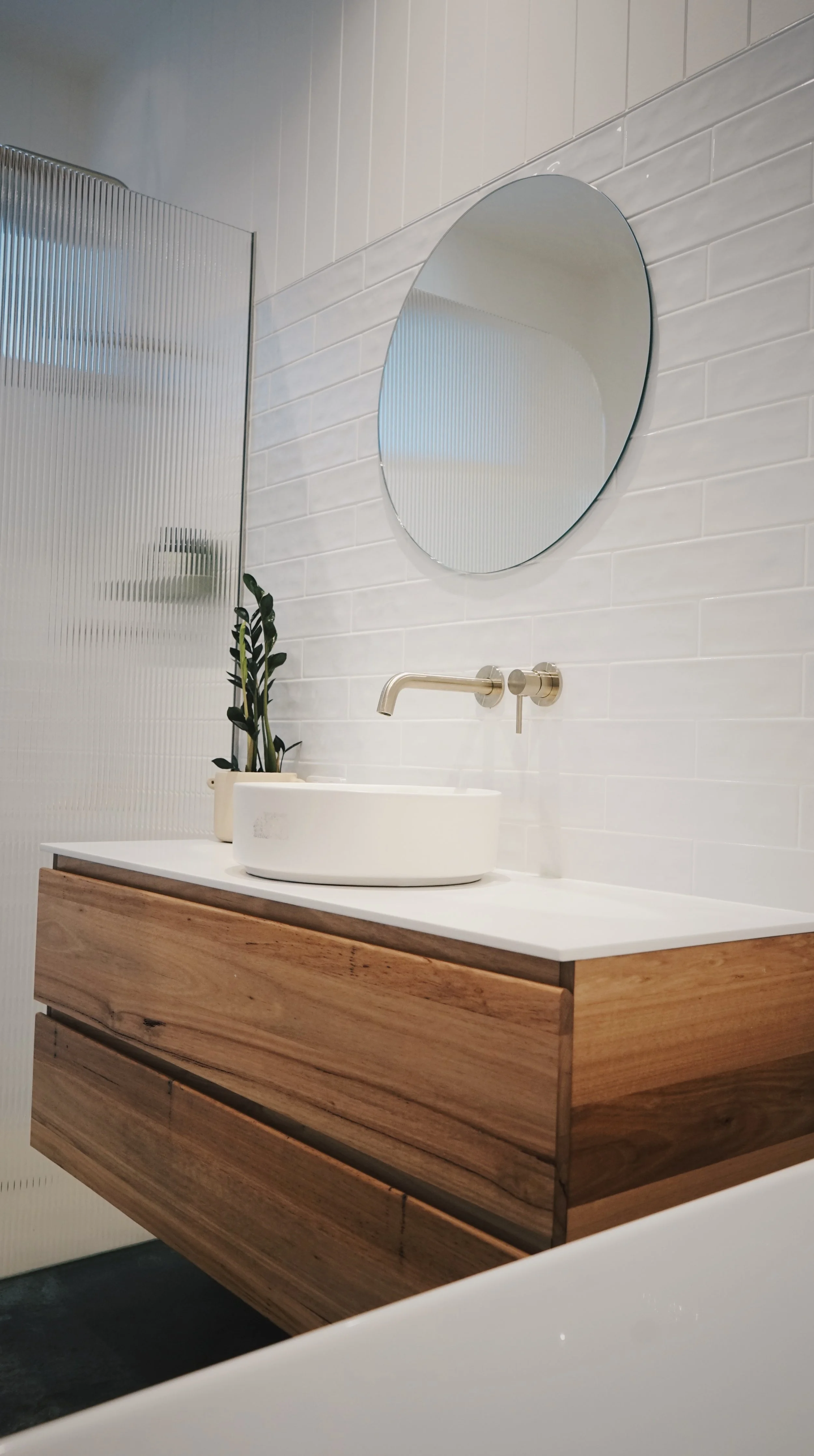 Modern bathroom with a wooden vanity, white countertop, vessel sink, wall-mounted faucet, round mirror, potted plant, and textured glass shower divider.