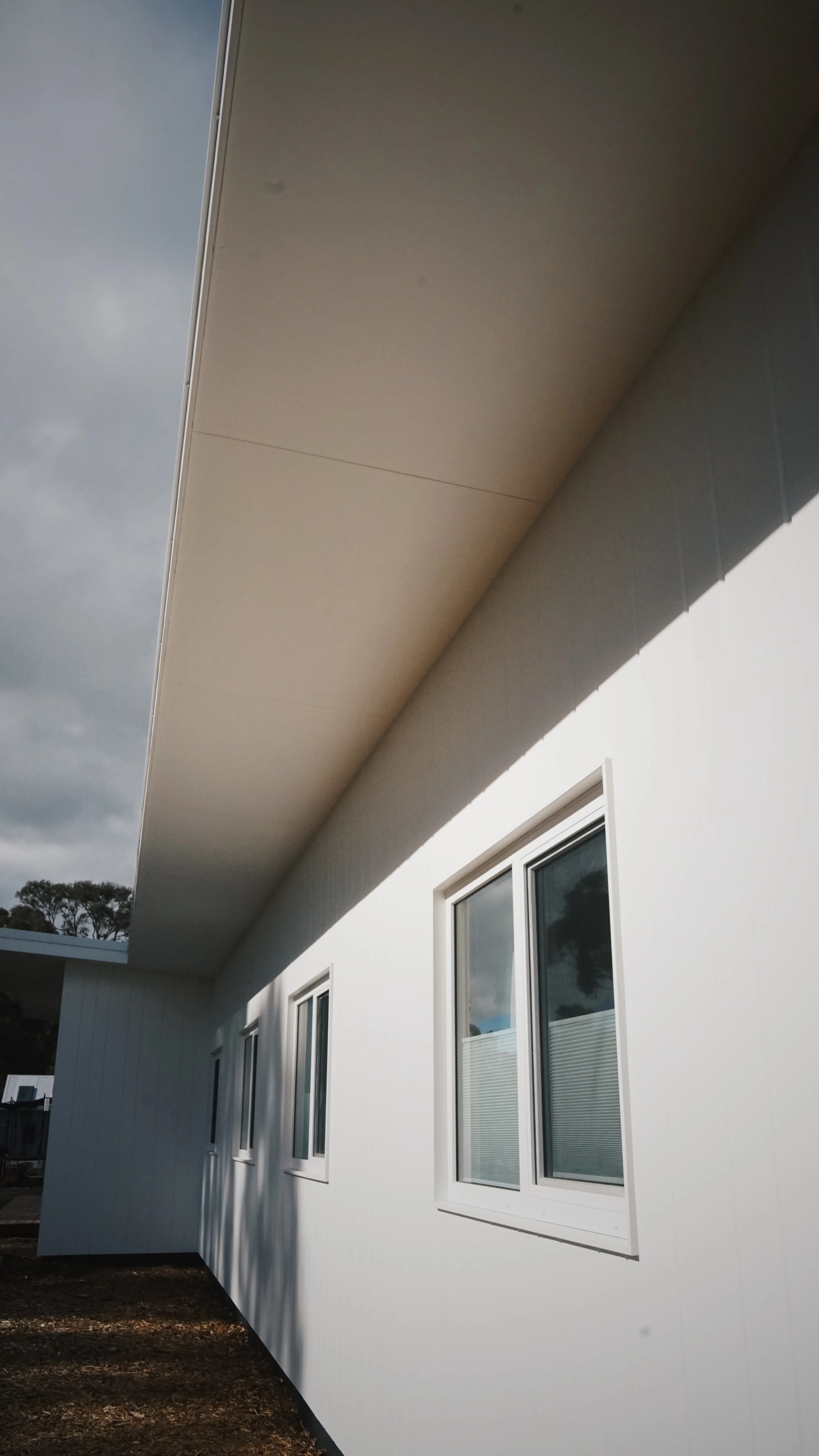 Side view of a modern white house with large rectangular windows and a protruding overhang or balcony under a cloudy sky.