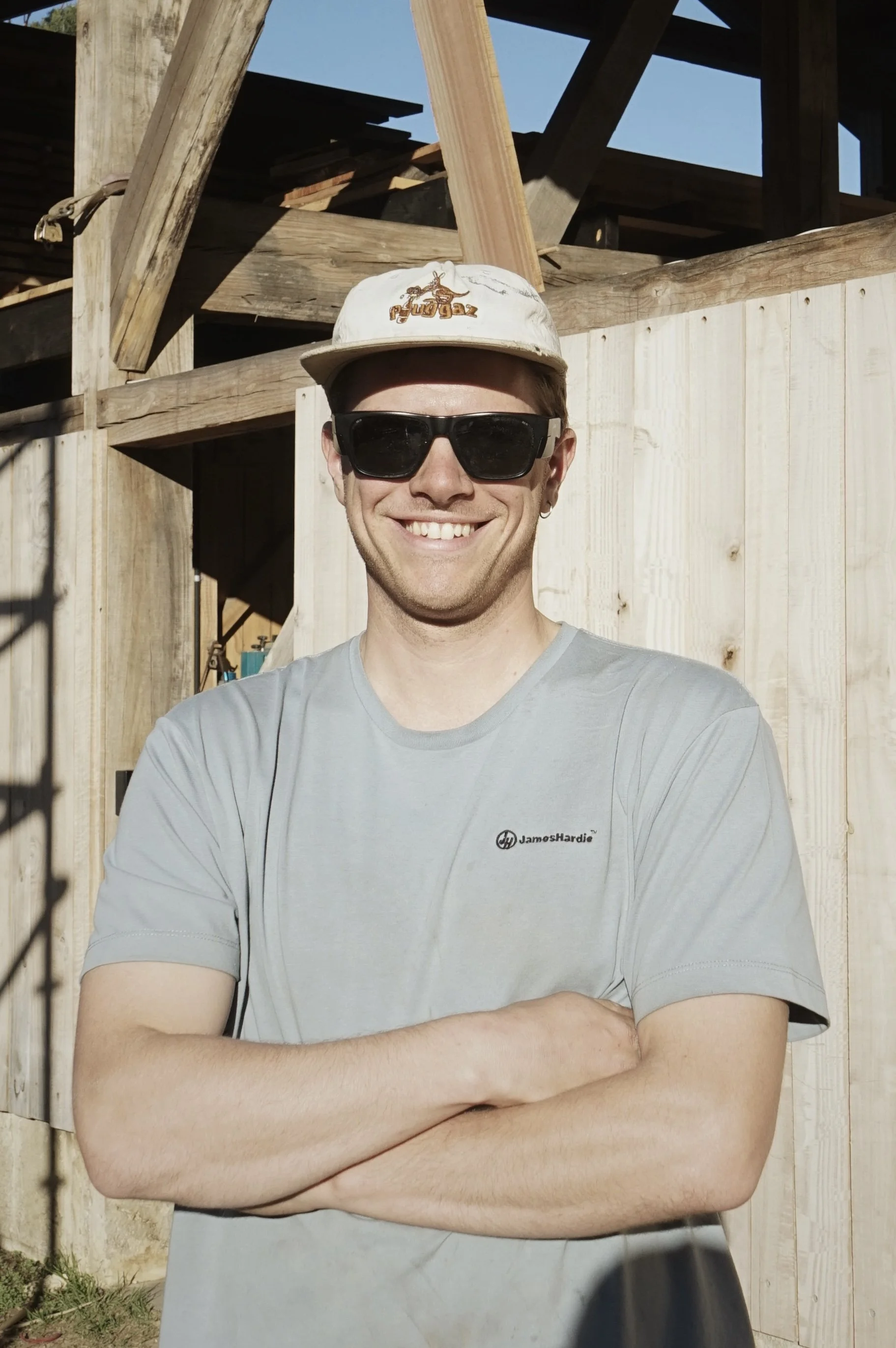 A smiling man with light skin, wearing a light gray T-shirt with the logo 'JamosHardie' and black sunglasses, standing outdoors in front of a wooden fence and a partially constructed wooden structure. He is also wearing a beige baseball cap with a logo that is not clearly visible.