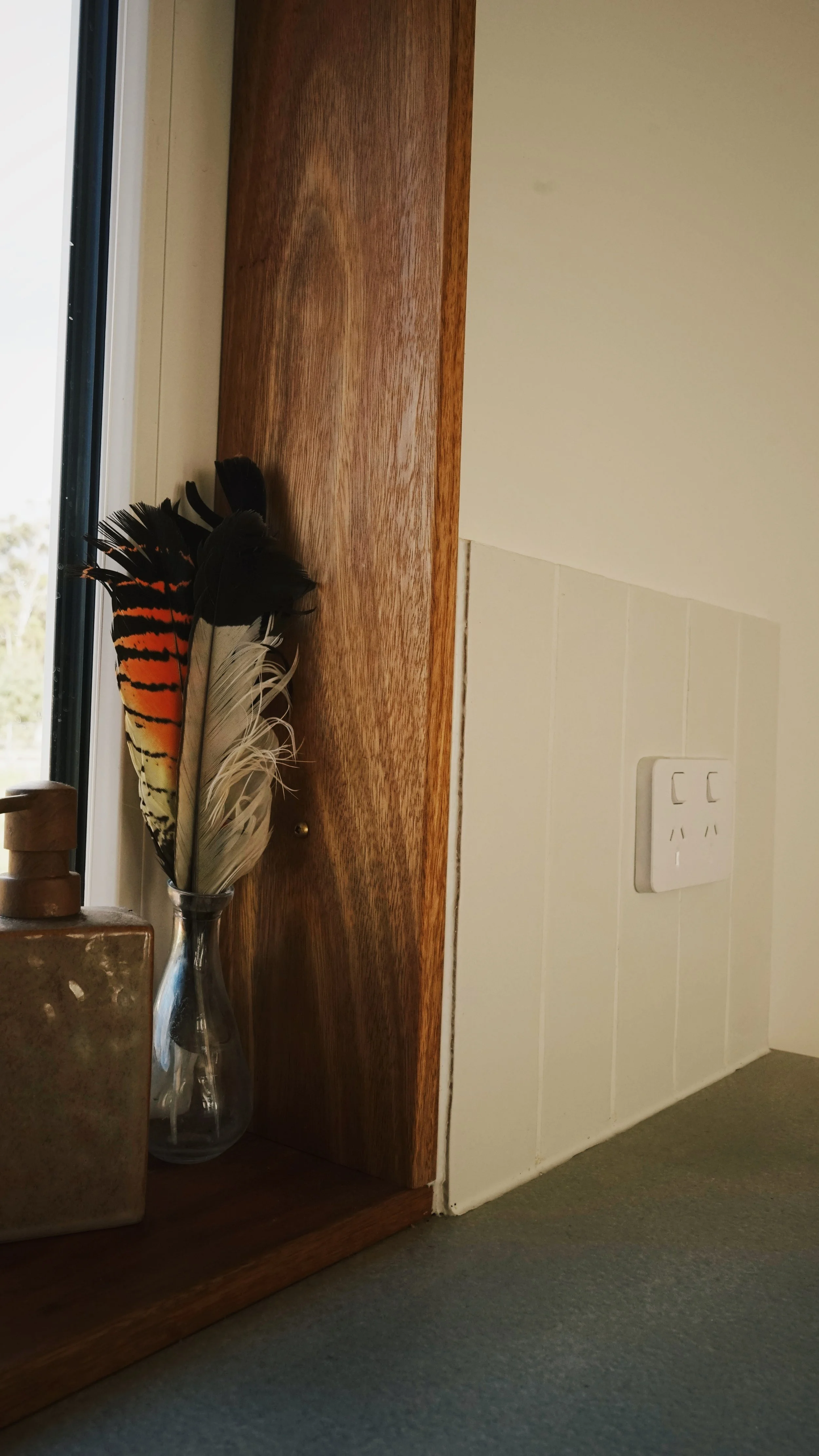 A wooden shelf with a glass vase holding colorful feathers next to a window, with a beige wall and electrical outlet nearby.